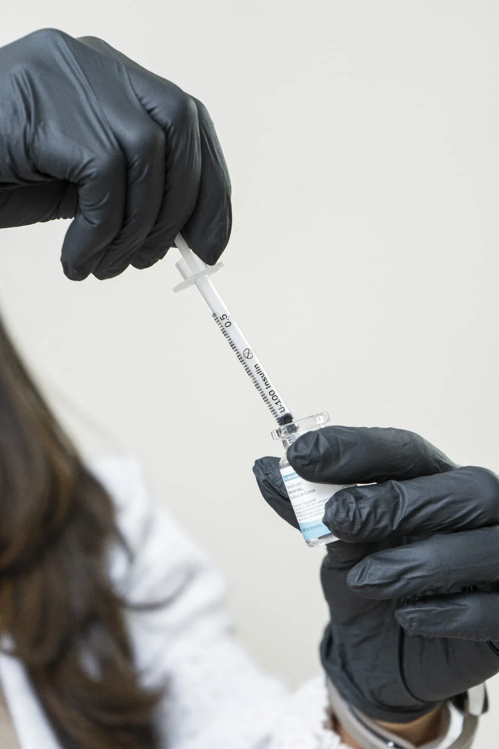 Personas con guantes negros preparando una dosis de vacuna con una jeringa y frasco, en un ambiente clínico.