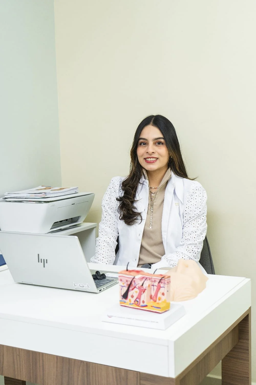 Mujer con blusa blanca y cabello largo sentado en escritorio de oficina con computadora portátil abierta y enviar documentos apilados