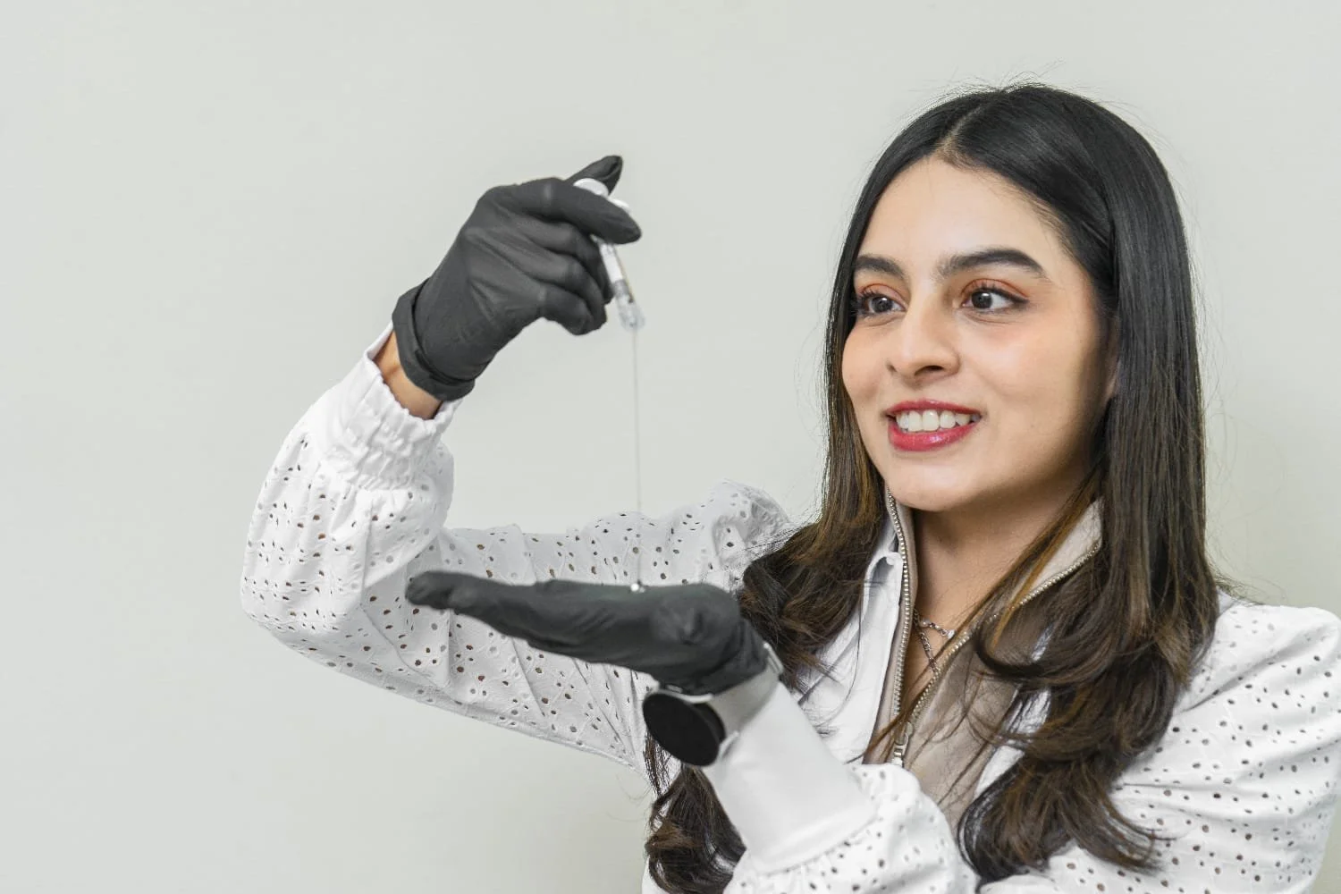 Joven mujer con bata de laboratorio y guantes negros sosteniendo una pipeta con líquido en actitud positiva.