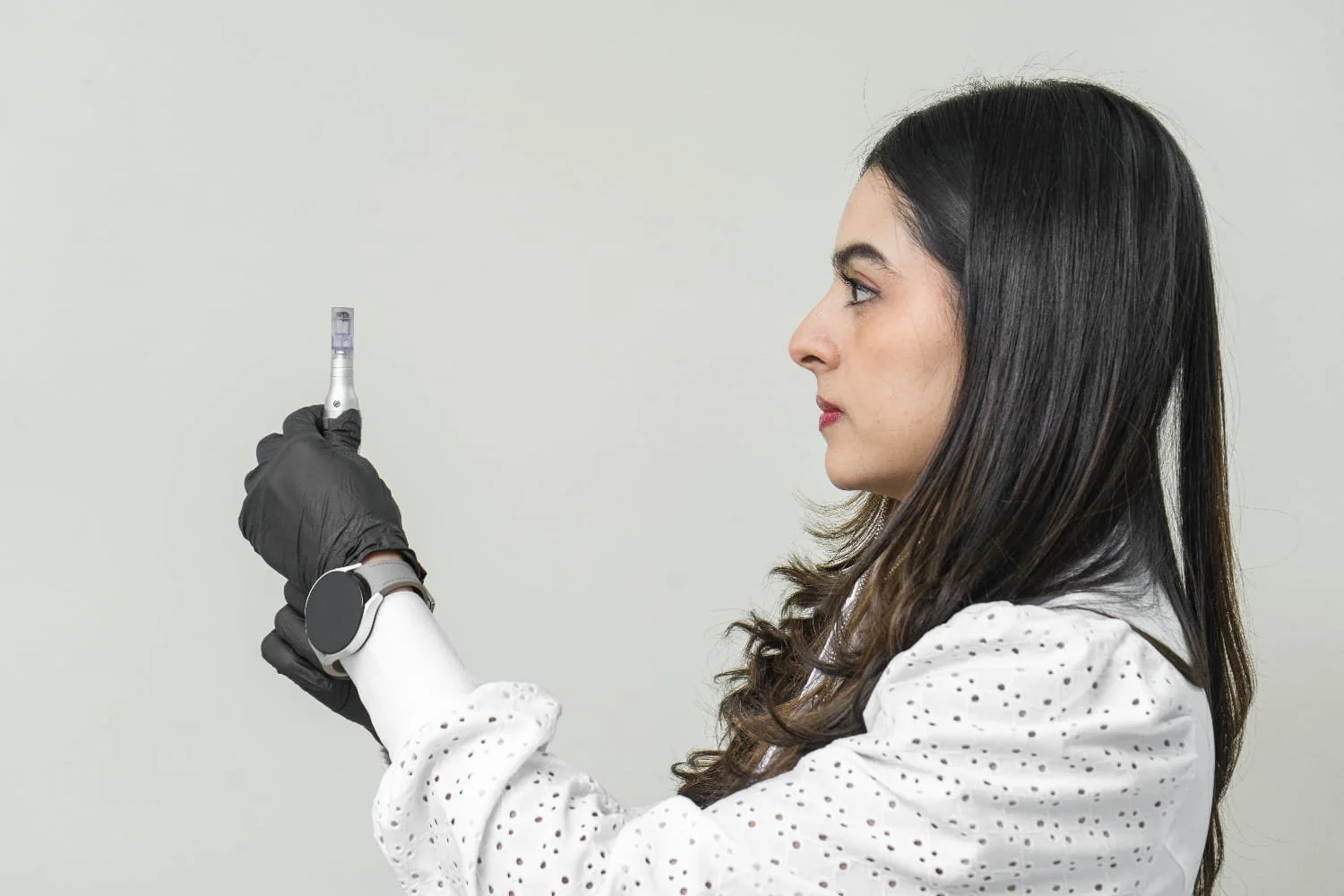 Mujer con bata blanca, sin maquillaje, con cabello largo y oscuro, sosteniendo un hisopo en la mano y usandos guantes negros, en un fondo gris claro.