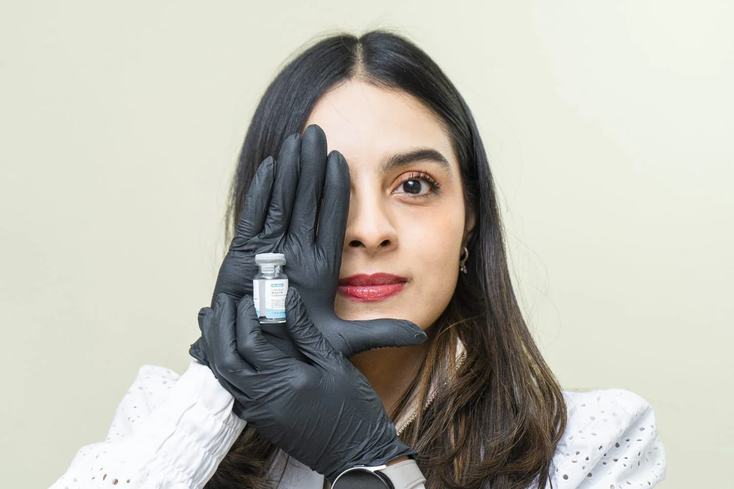 Mujer con cabello largo y piel clara sosteniendo una vacuna con guantes negros frente a su rostro de perfil, con un fondo neutral.