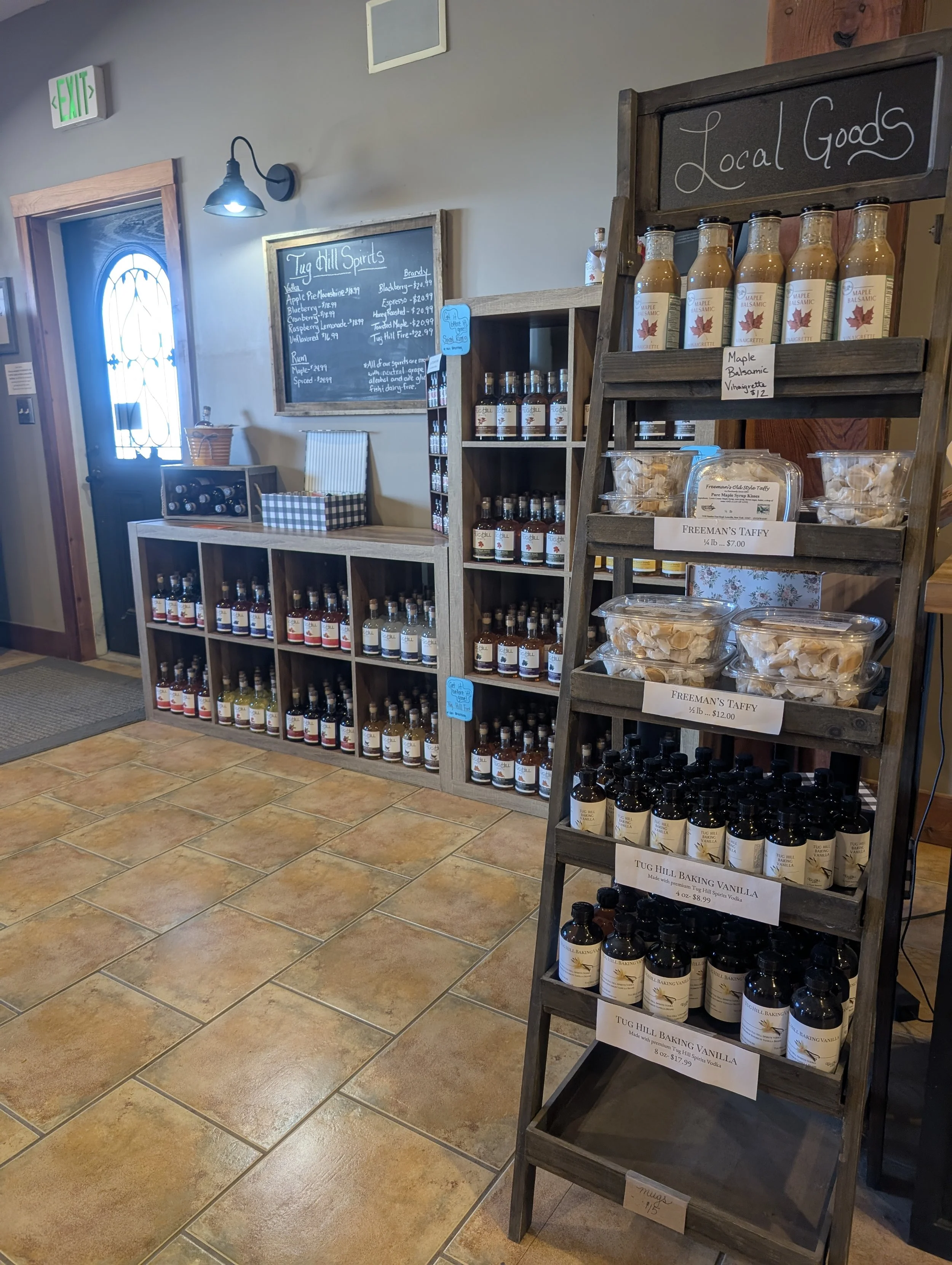 Interior of a store with shelves containing bottles of flavored alcohol and a chalkboard menu listing liquors including Tug Hill Spirits and Freeman's Taffy, near a door with a glass window and a green illuminated exit sign.