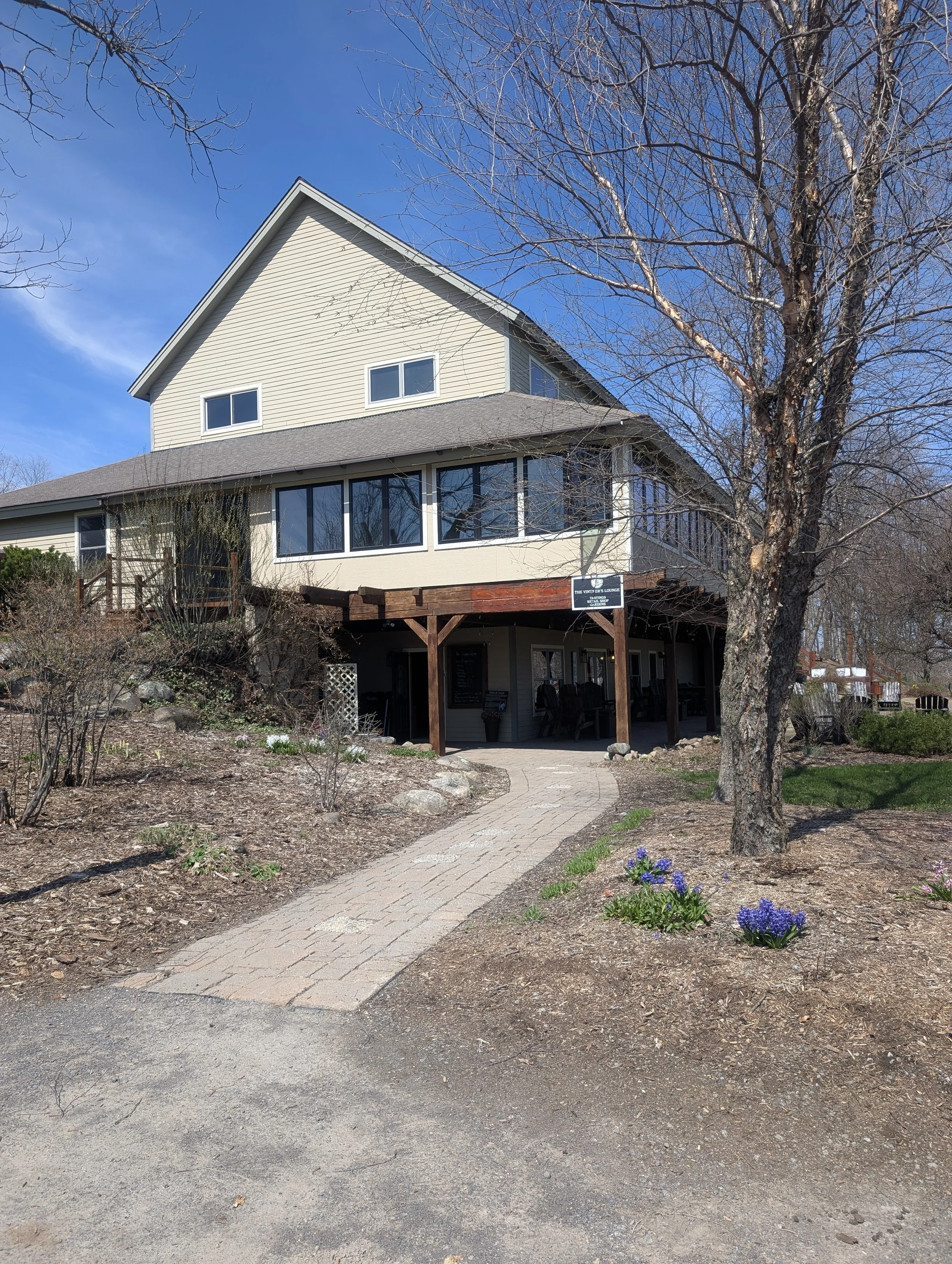View of a two-story house with beige siding, large windows, and a screened porch, surrounded by leafless trees and a garden with purple flowers, under a clear blue sky.