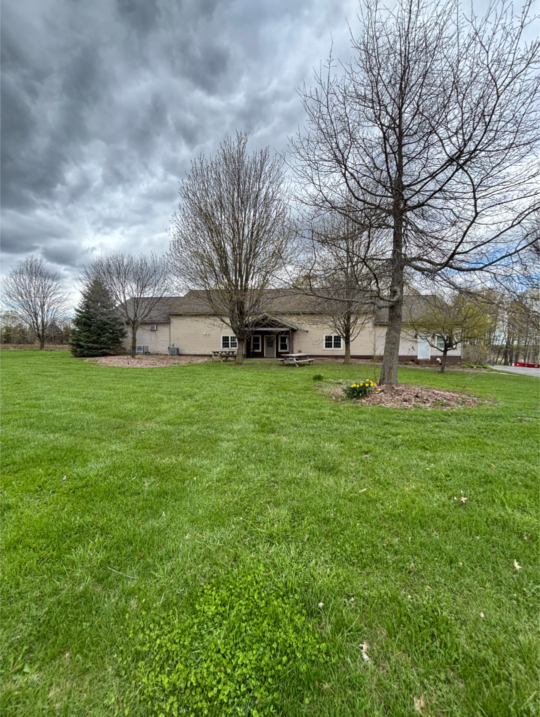 A grassy yard with a house in the background. The house has a porch with picnic tables, some trees without leaves, and a cloudy sky overhead.