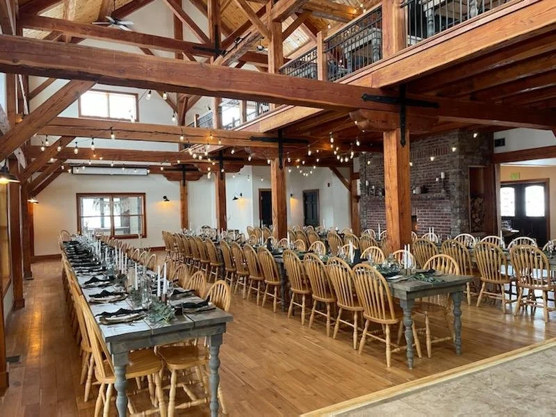Long banquet tables set for a large gathering in a rustic event hall with exposed wooden beams and string lights.