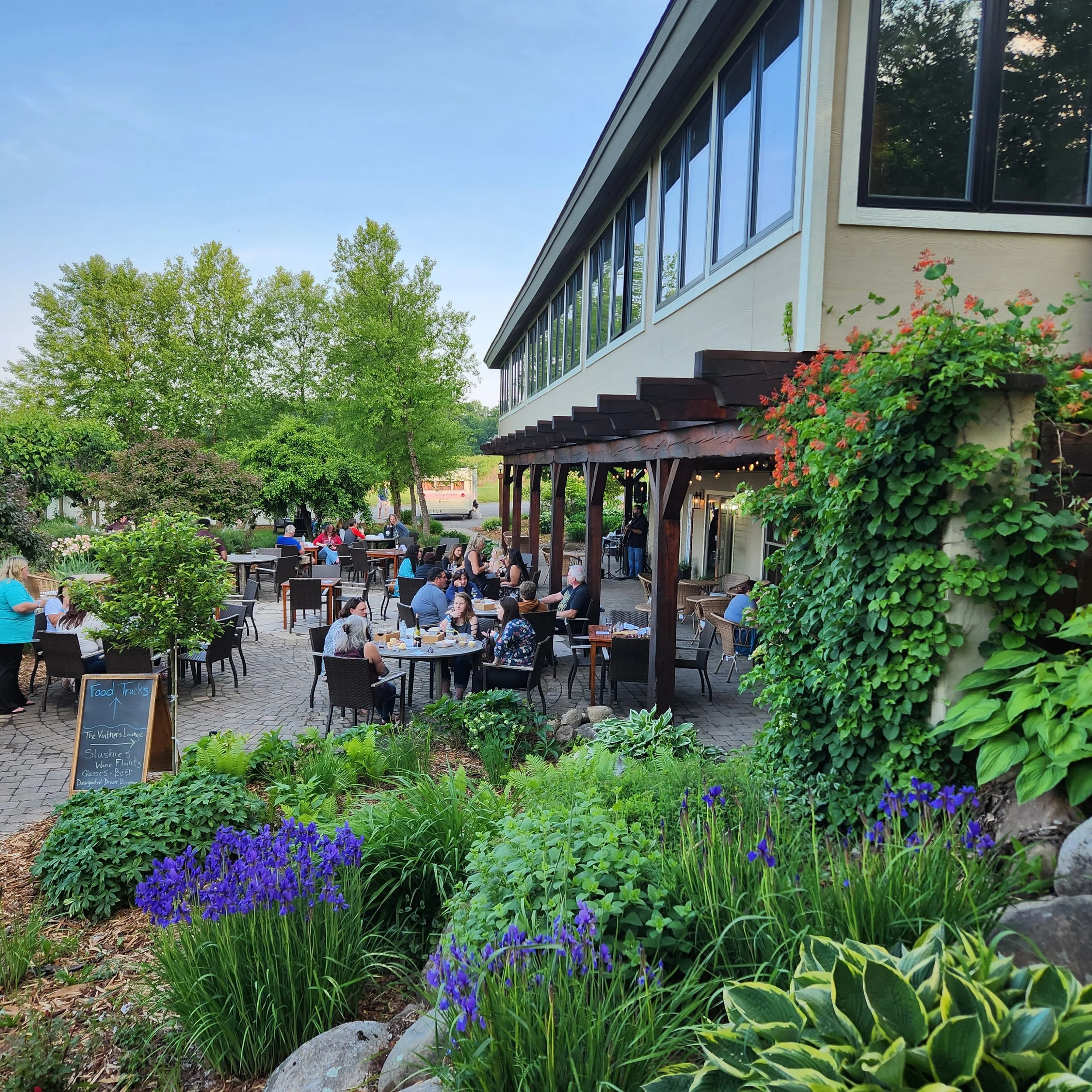 An outdoor patio area with people sitting at tables, a building with large windows on the right, lush green trees, various plants and purple flowers in the foreground, and a blue sky overhead.