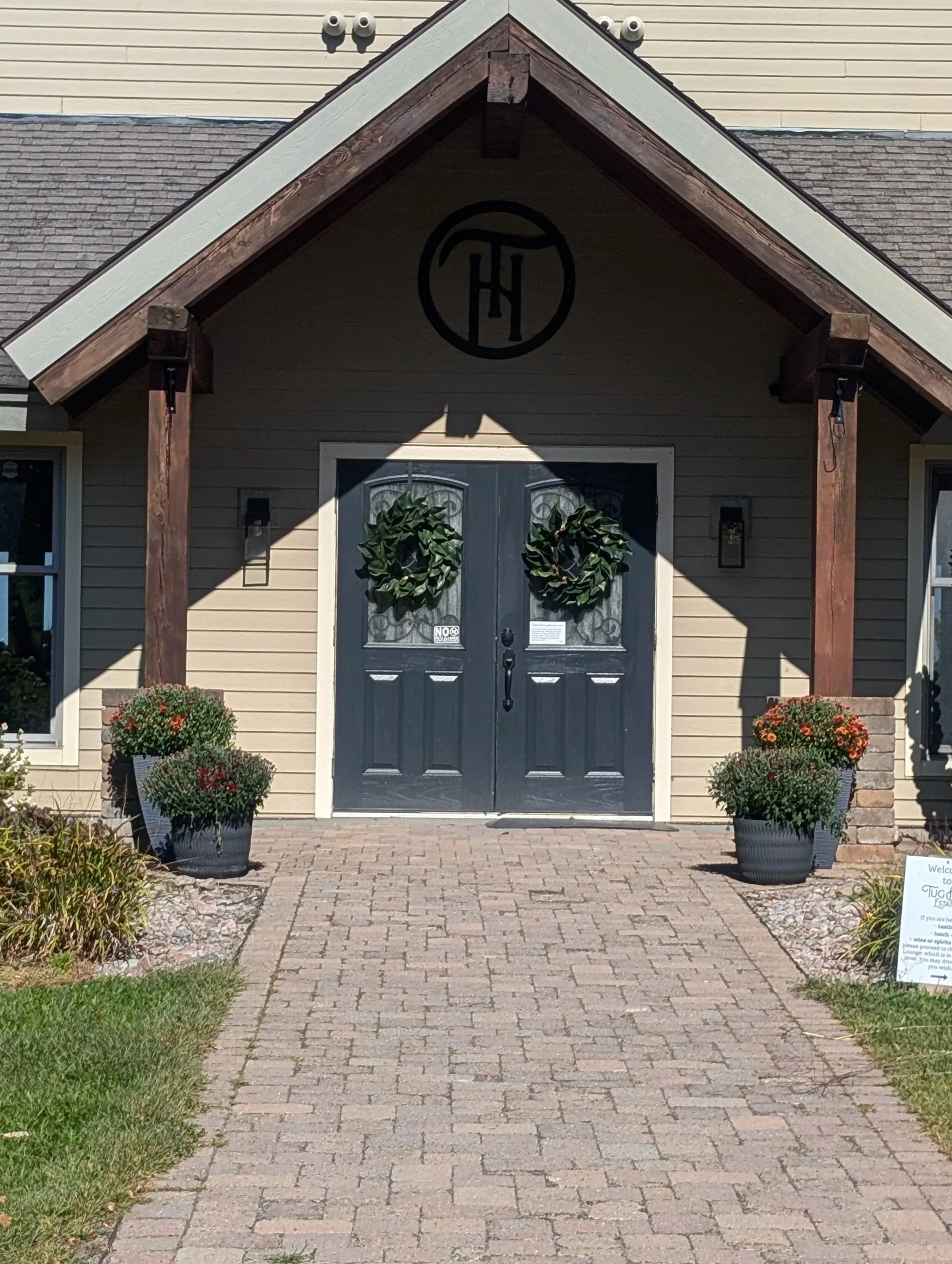 Front entrance of a house with a double door decorated with wreaths, flanked by two potted plants, a brick pathway, and a sign on the right side.