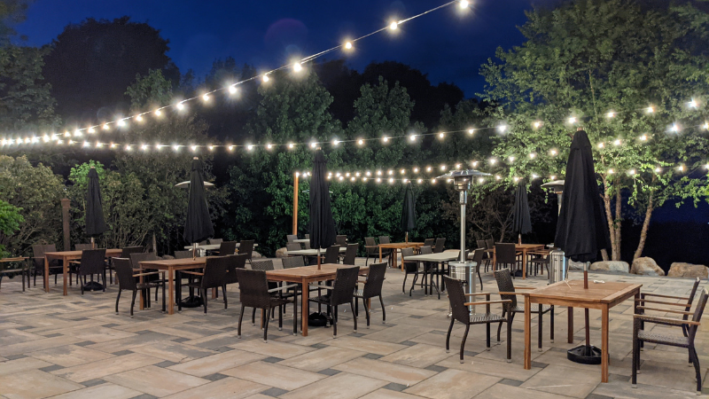 Outdoor patio at night with string lights overhead, wooden tables, and black umbrellas among trees.