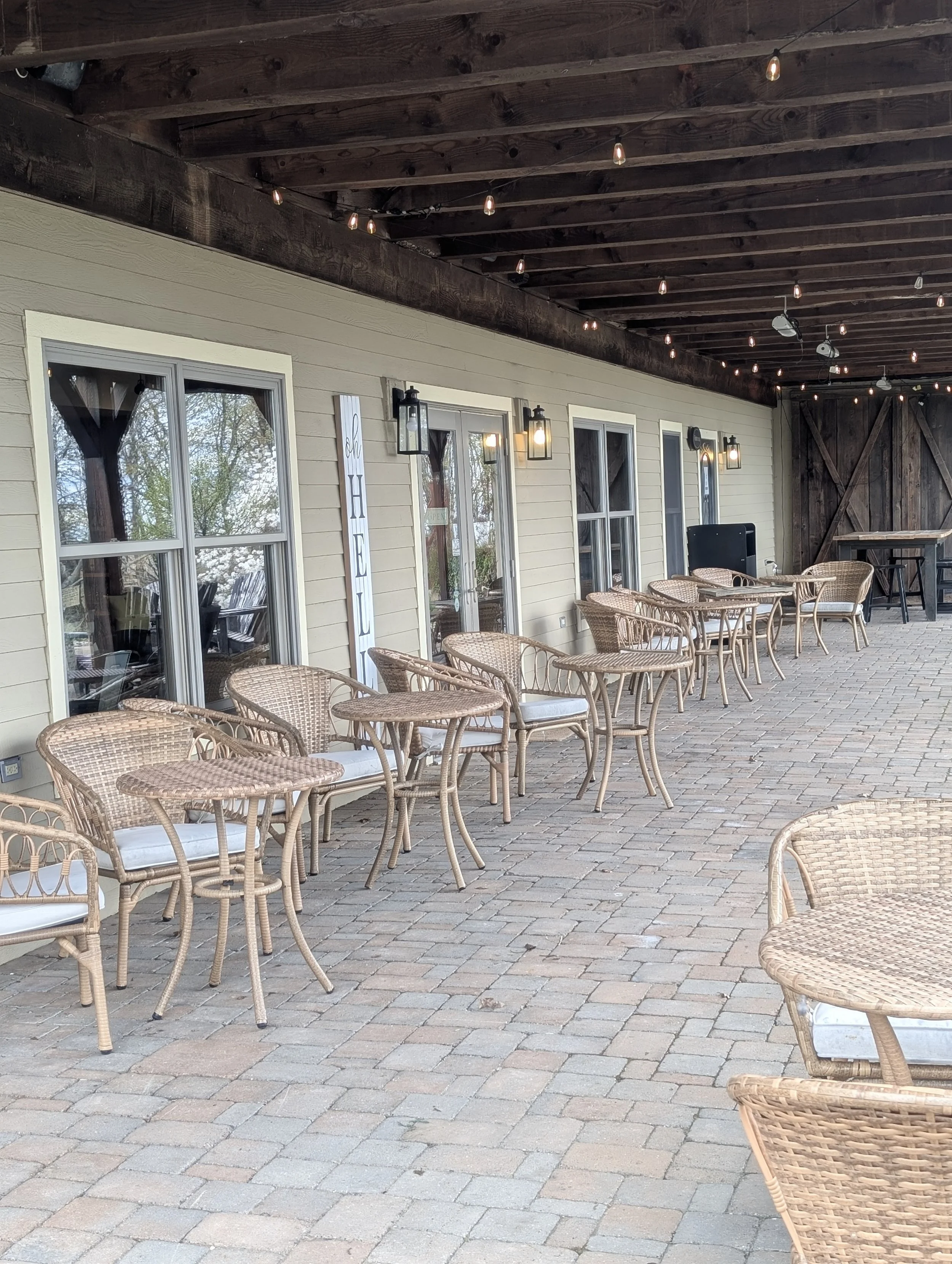 Outdoor patio area with wicker chairs and tables, multiple windows on the house, string lights hanging from a dark wooden ceiling, and a sign that reads "Oh Hello".