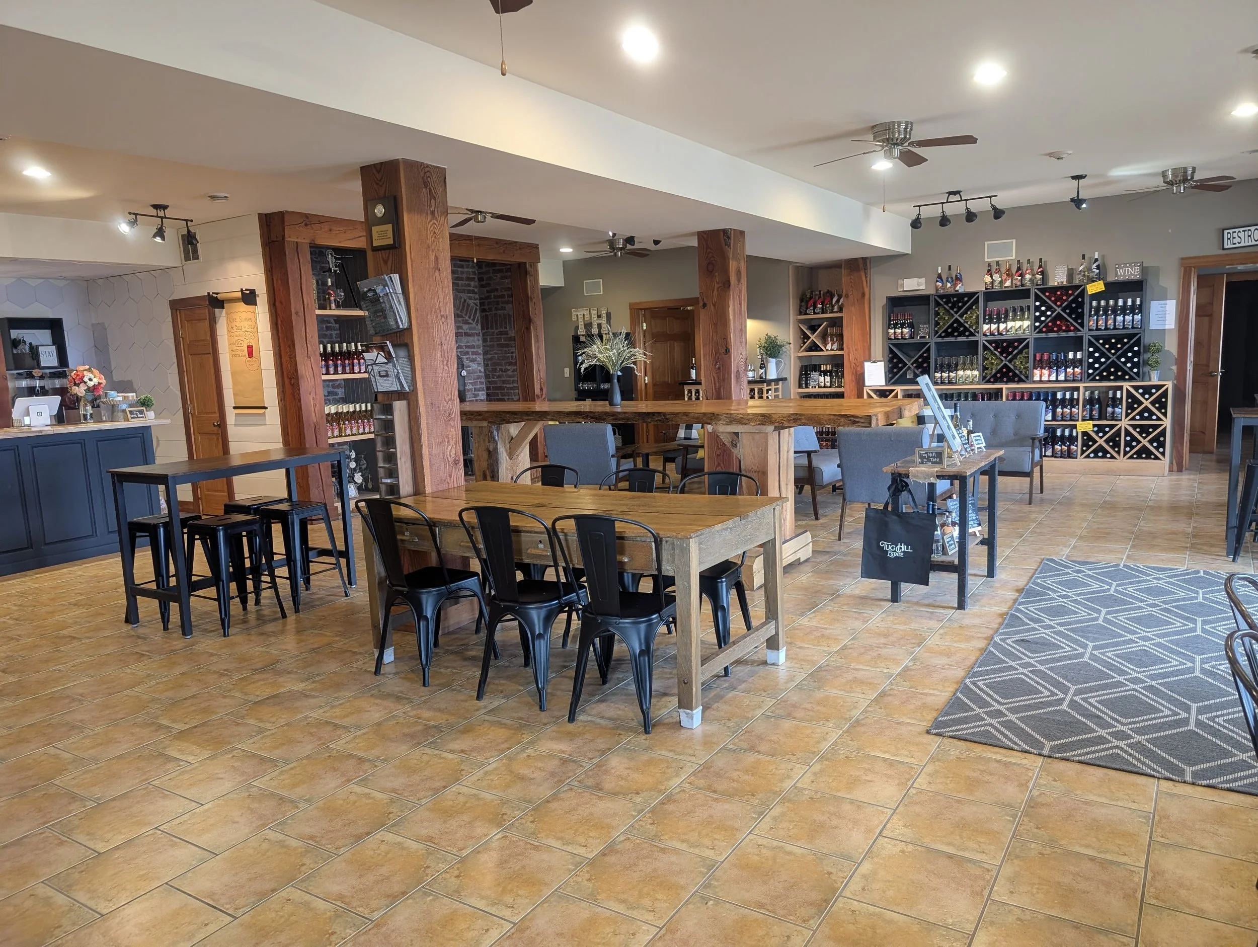 Interior of a restaurant or café with wooden tables, black chairs, wine racks, and decorative plants, featuring a modern rustic design with plaid rug and ceiling fans.