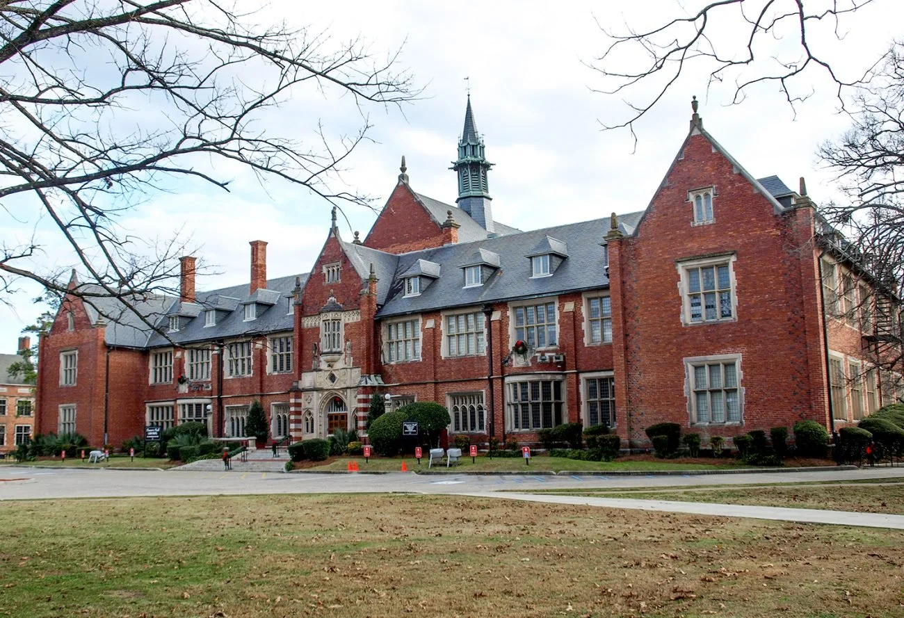 Large historic brick building with gothic architecture, multiple windows, and a central steeple, surrounded by leafless trees and a grassy area in the foreground.