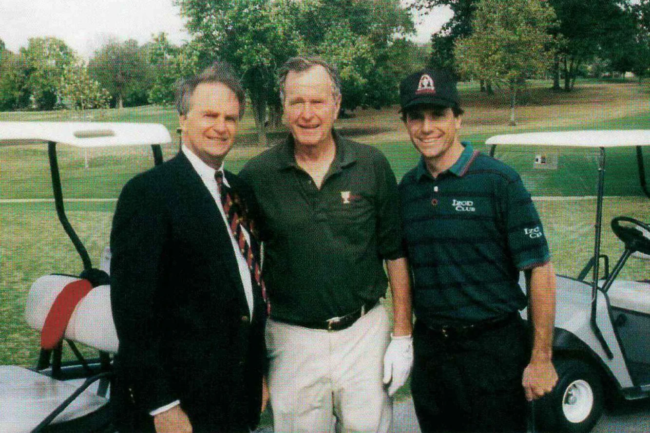 Three men standing on a golf course, smiling, with golf carts in the background.