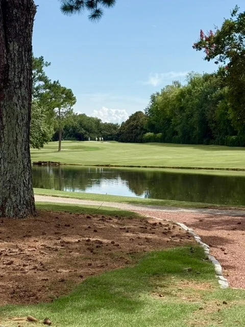 Scenic view of a golf course with a pond, tall trees, a well-maintained green, and a clear blue sky.
