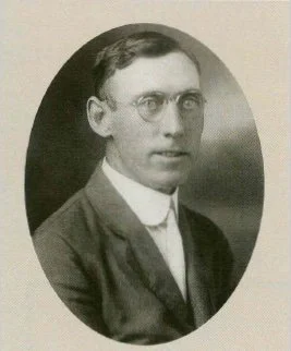 A vintage black-and-white portrait of a young man with glasses, wearing a suit and tie, in an oval frame.