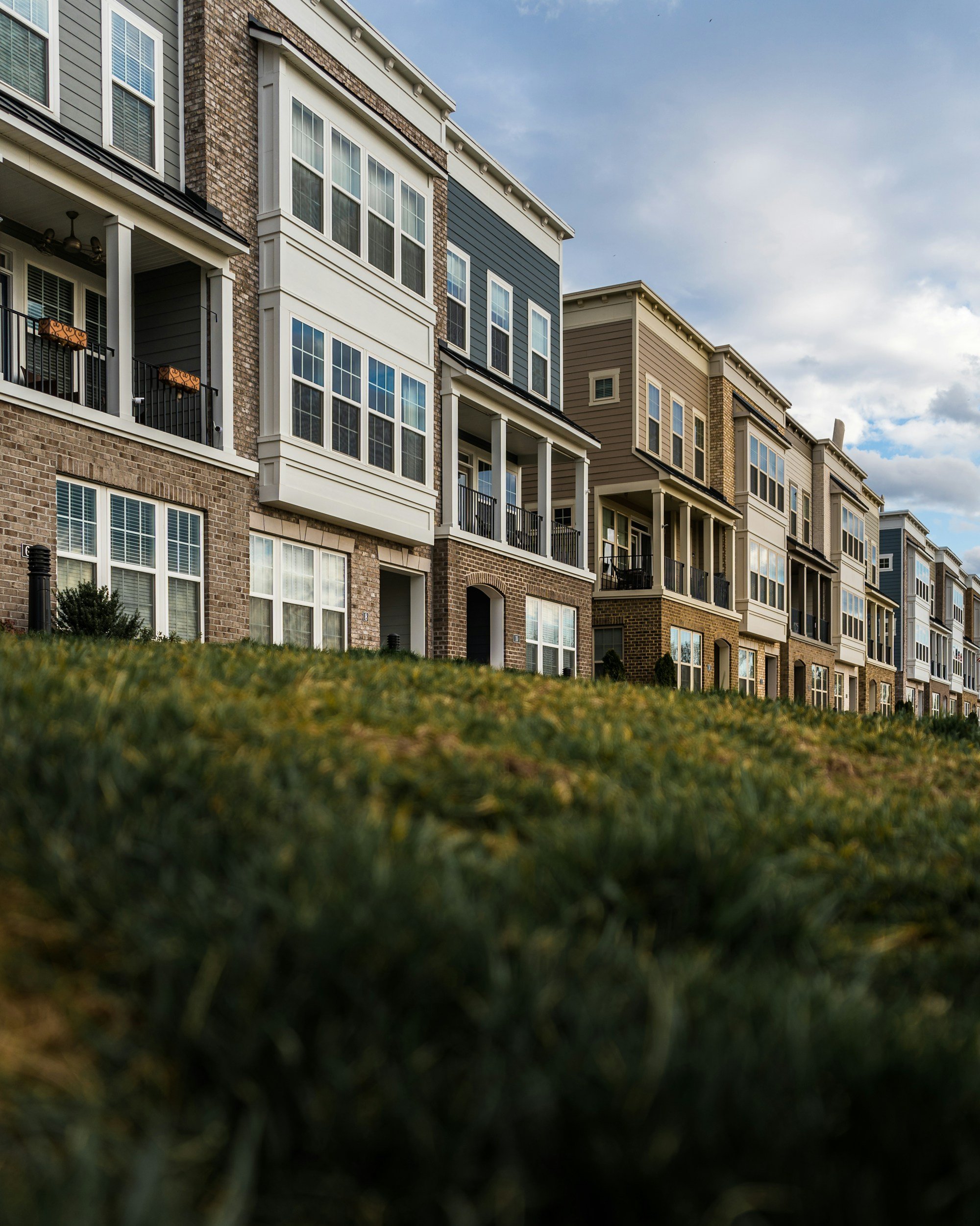 A row of modern multi-story apartment buildings with various exterior colors and designs, viewed from a low angle with front yards and a cloudy sky in the background.