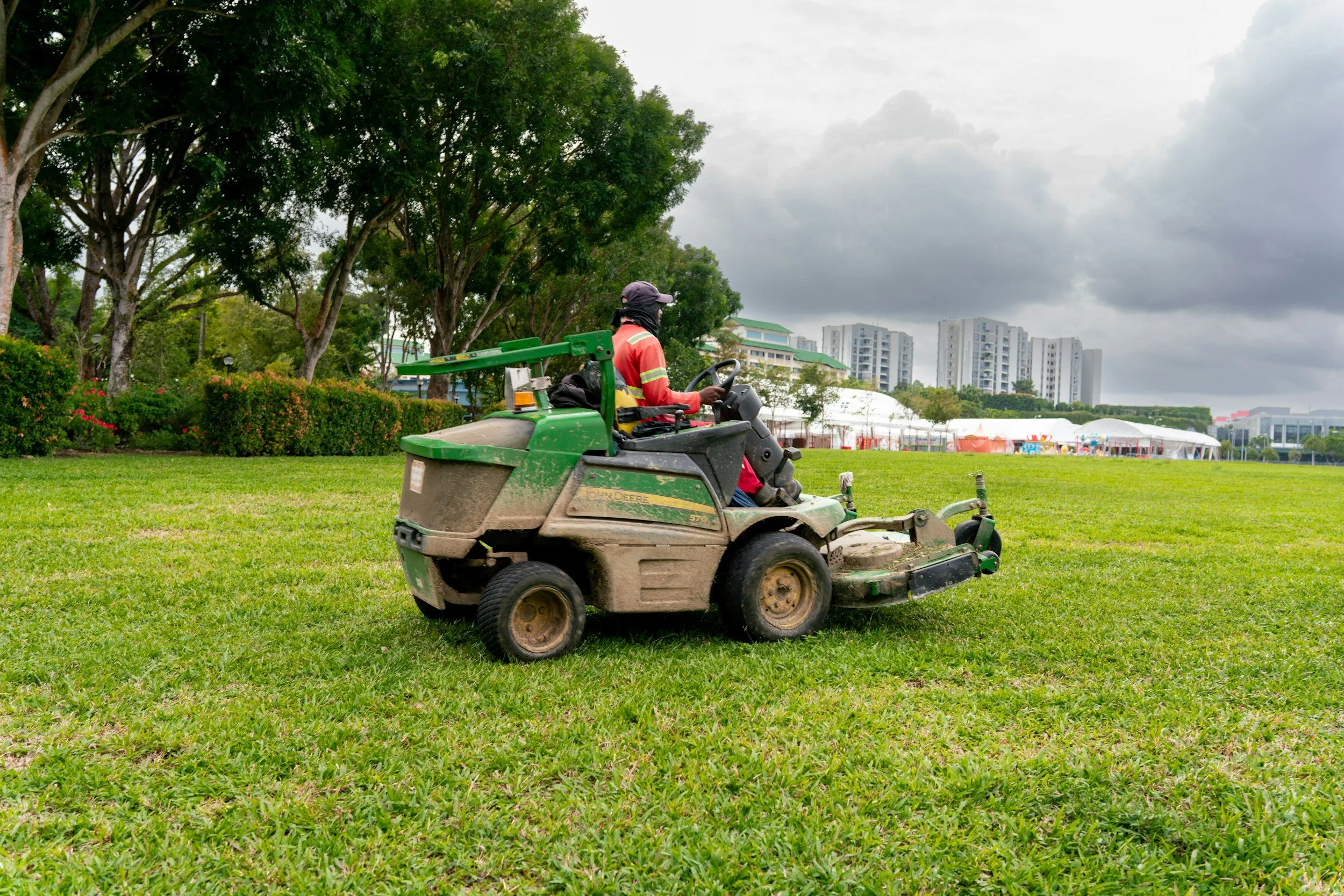 A person operating a John Deere riding lawn mower on a grassy field with trees and buildings in the background, under a cloudy sky.