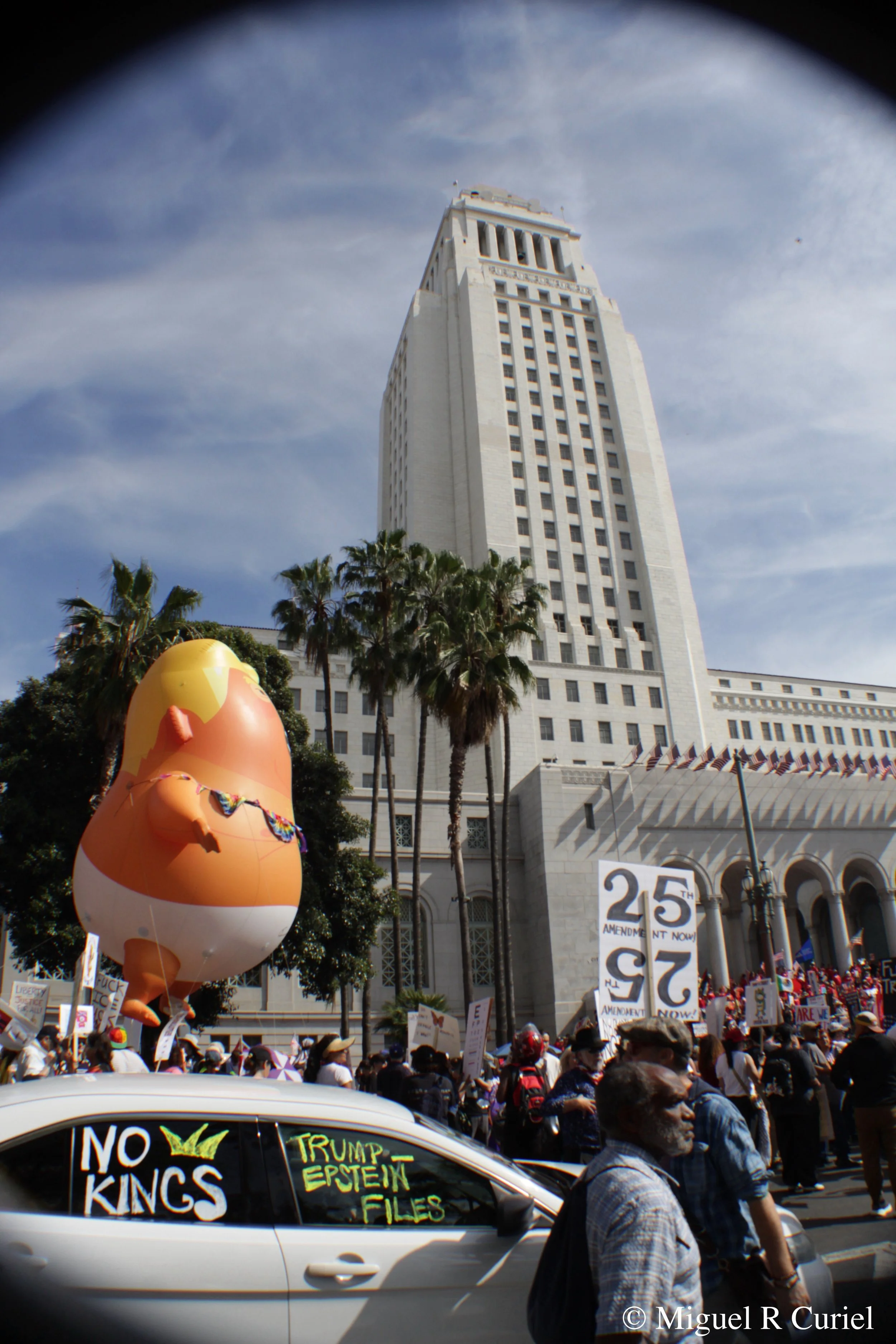 A protest in front of the Los Angeles City Hall, with a large inflatable figure of a baby Donald Trump in front of palm trees.  'NO KINGS' and 'TRUMP ESPIONAGE FILES' are painted on a car window in the foreground. 