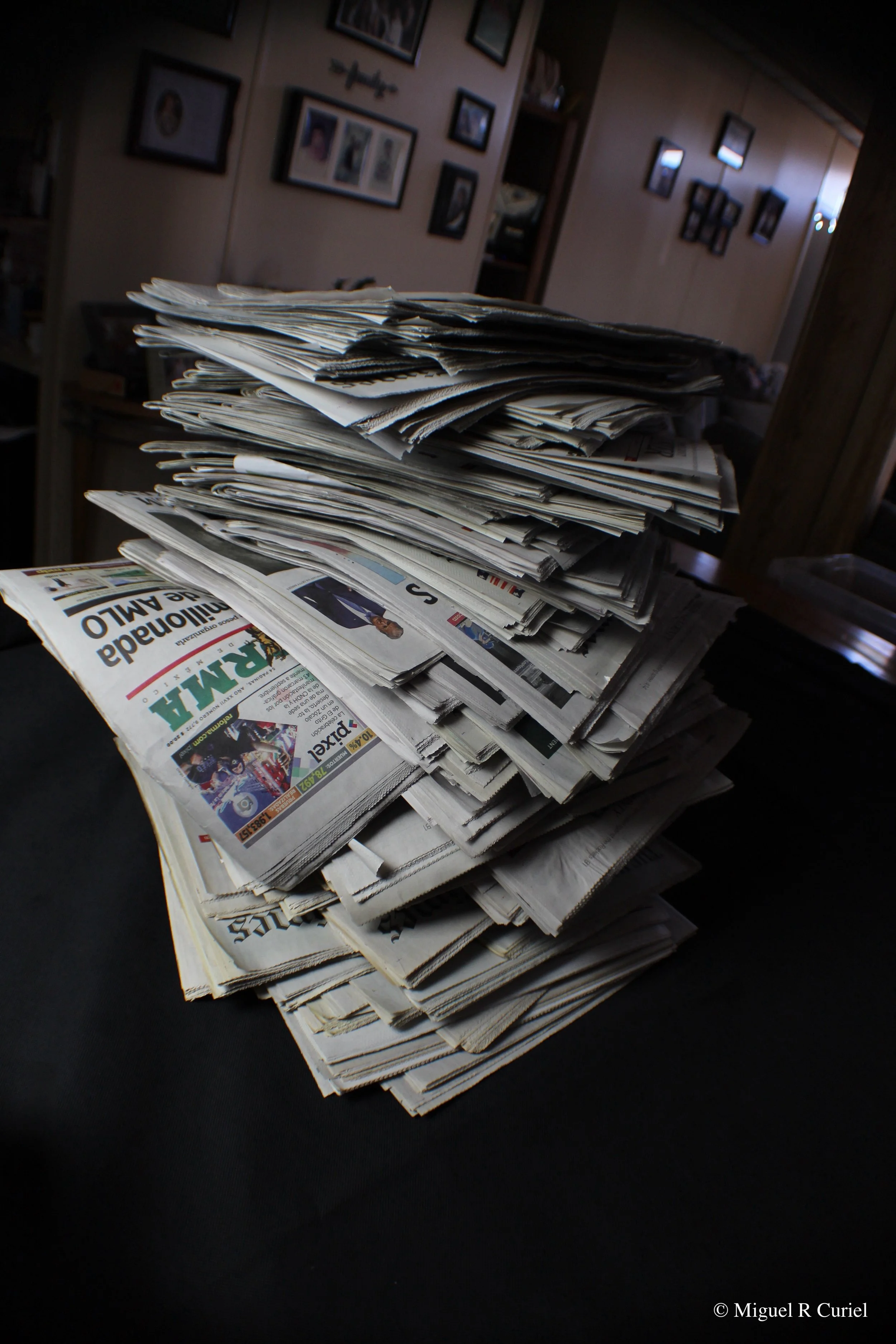 A large pile of newspapers stacked on a dark surface, with a blurred interior background of a room decorated with framed pictures on the walls.