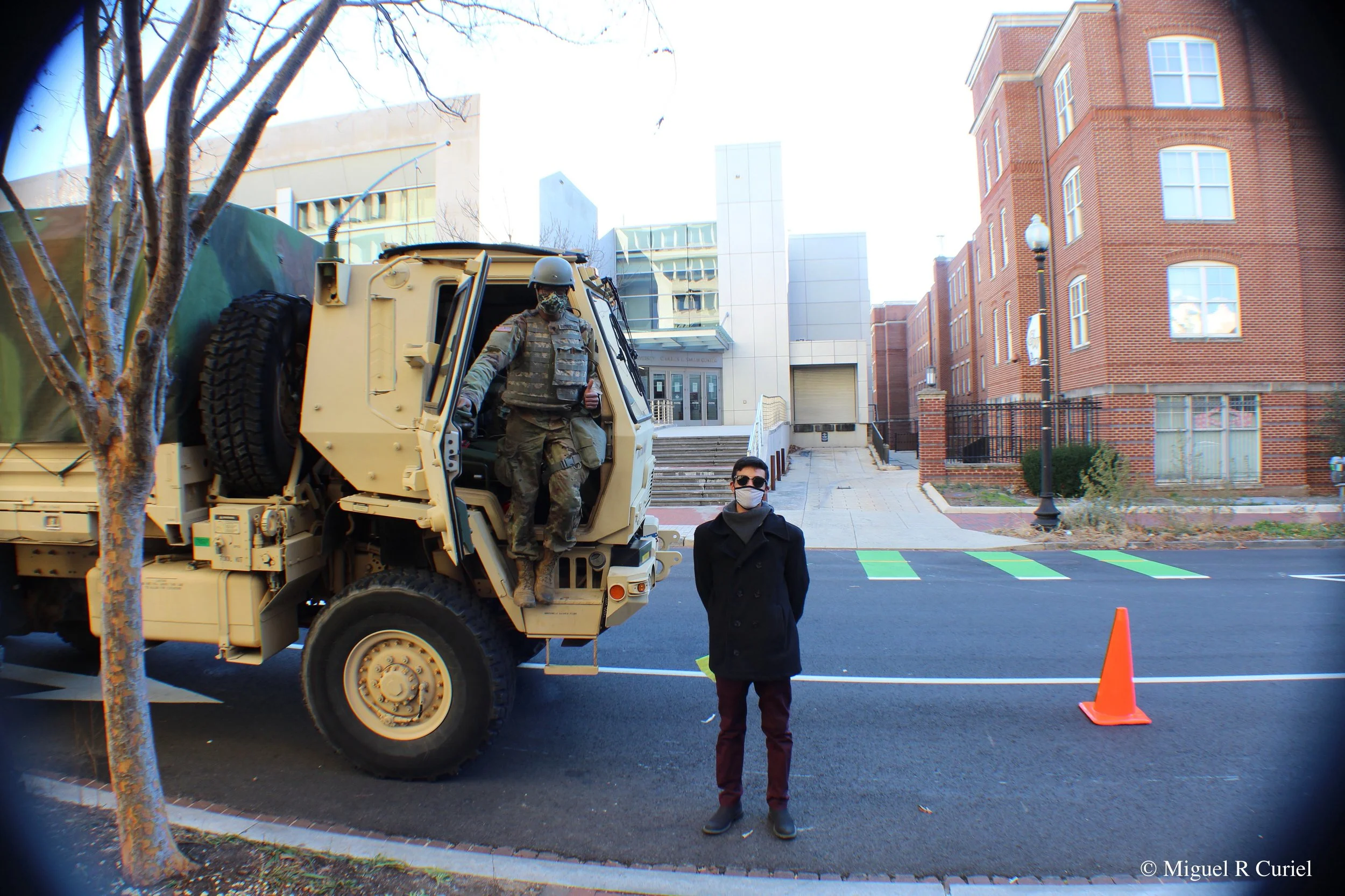 Miguel Curiel in civilian clothing and a face mask stands in the streets of Washington DC in front of a military vehicle with a member of the National Guard standing at the door way.