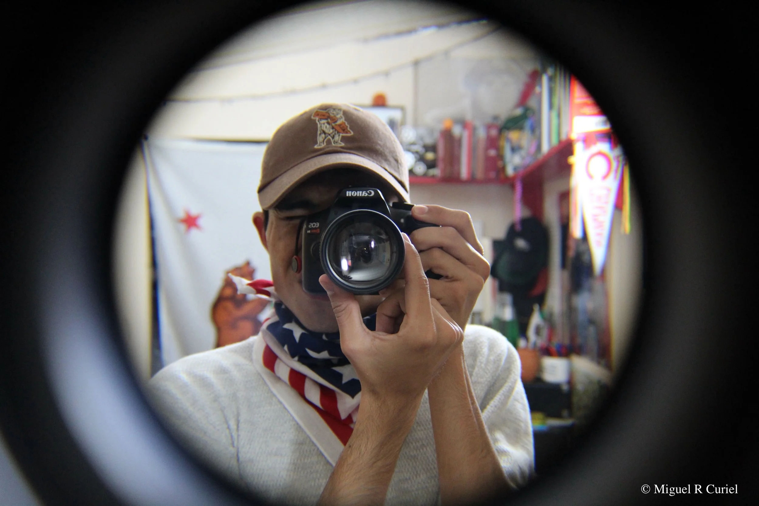 Miguel Curiel taking a selfie with a camera, wearing a brown hat with a patch of the California Bear hugging the State.