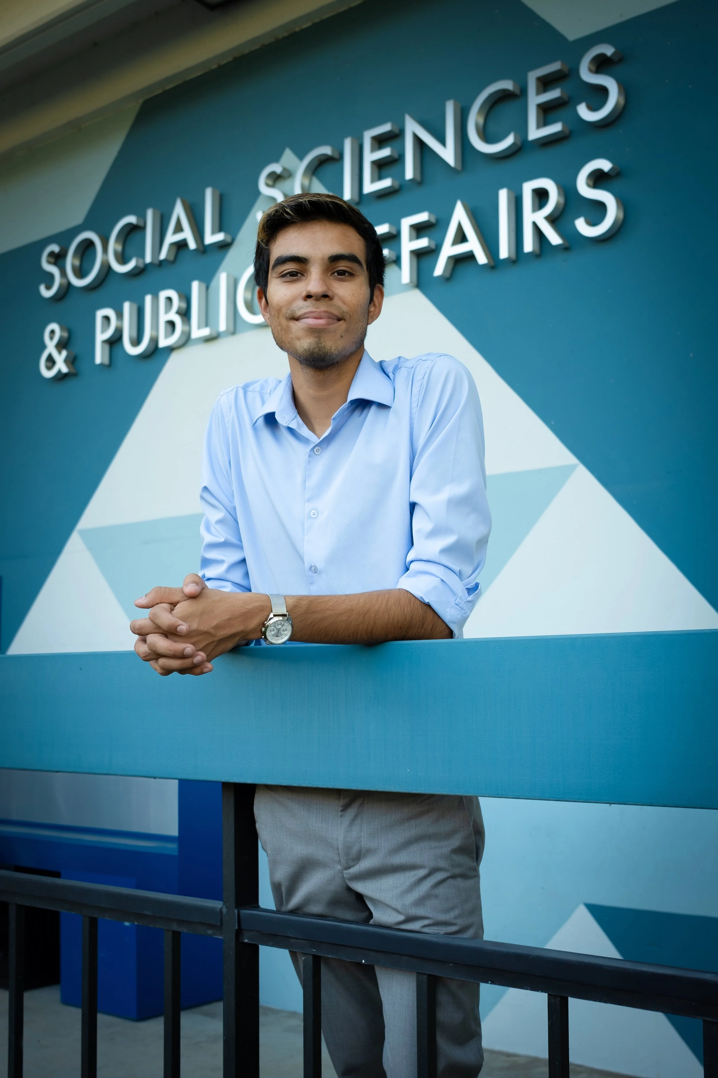 Miguel Curiel in a light blue shirt leaning on a blue barrier outside a building with a sign that reads 'Social Sciences & Public Fairs'.