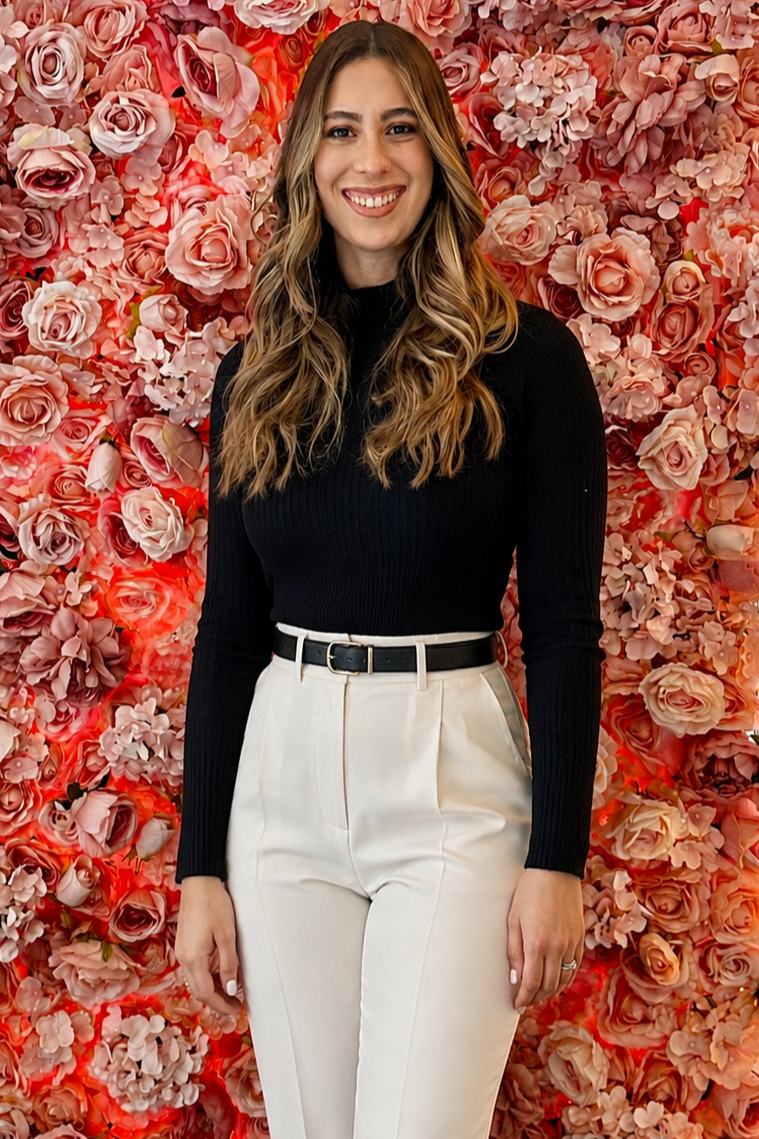 A therapist with long wavy blonde hair that specializes in treating maternal mental health smiling, wearing a black long-sleeve top and high-waisted white pants with a black belt, standing in front of a wall covered in pink and peach roses.