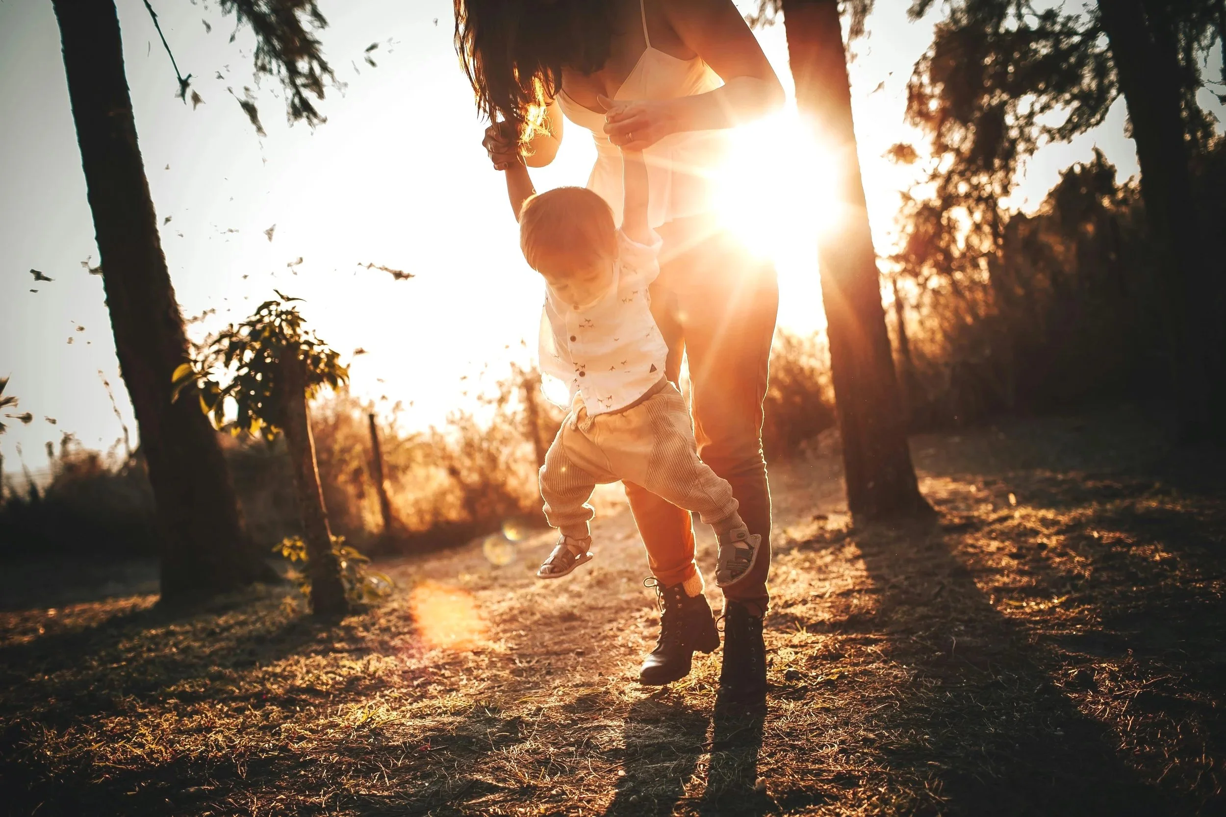 A woman lifting a young child in a park in Middletown, NY during sunset, surrounded by trees, with warm sunlight creating a glowing backlight.