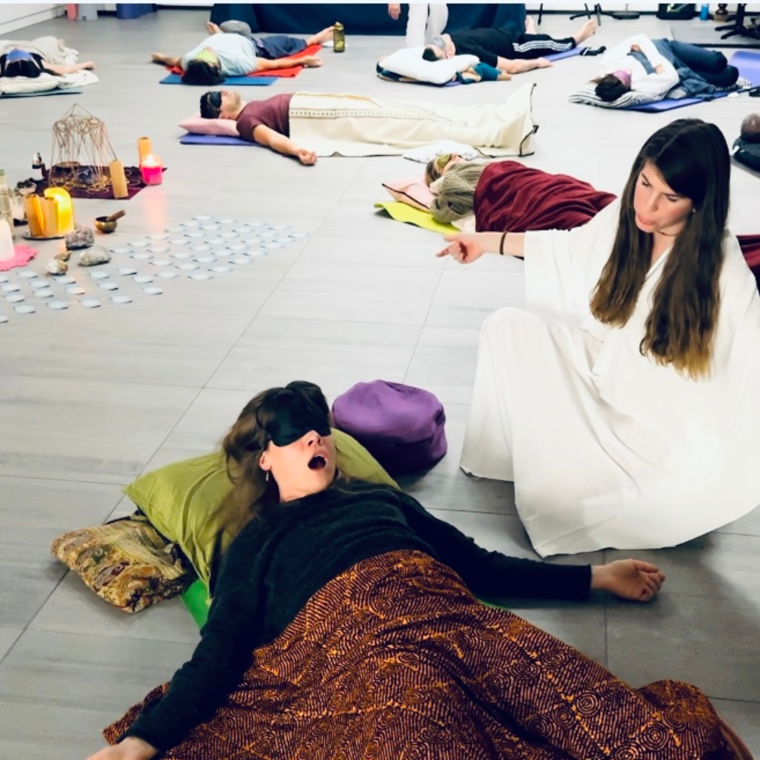 People participating in a guided meditation or yoga session, lying on yoga mats on the floor, with candles and objects arranged in a decorative circle nearby.
