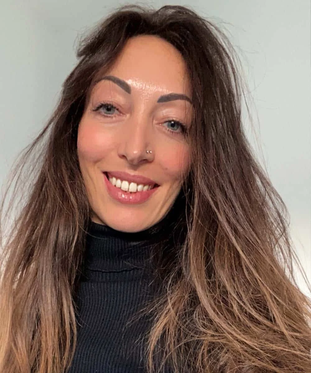 A woman with long, wavy brown hair and light blue eyes smiling, wearing a black turtleneck against a plain light-colored background.