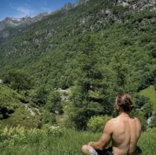 A shirtless person sitting cross-legged outdoors, overlooking a lush green valley surrounded by mountains and trees.
