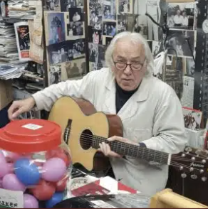 An elderly man playing an acoustic guitar in a cluttered room filled with photographs and miscellaneous items.