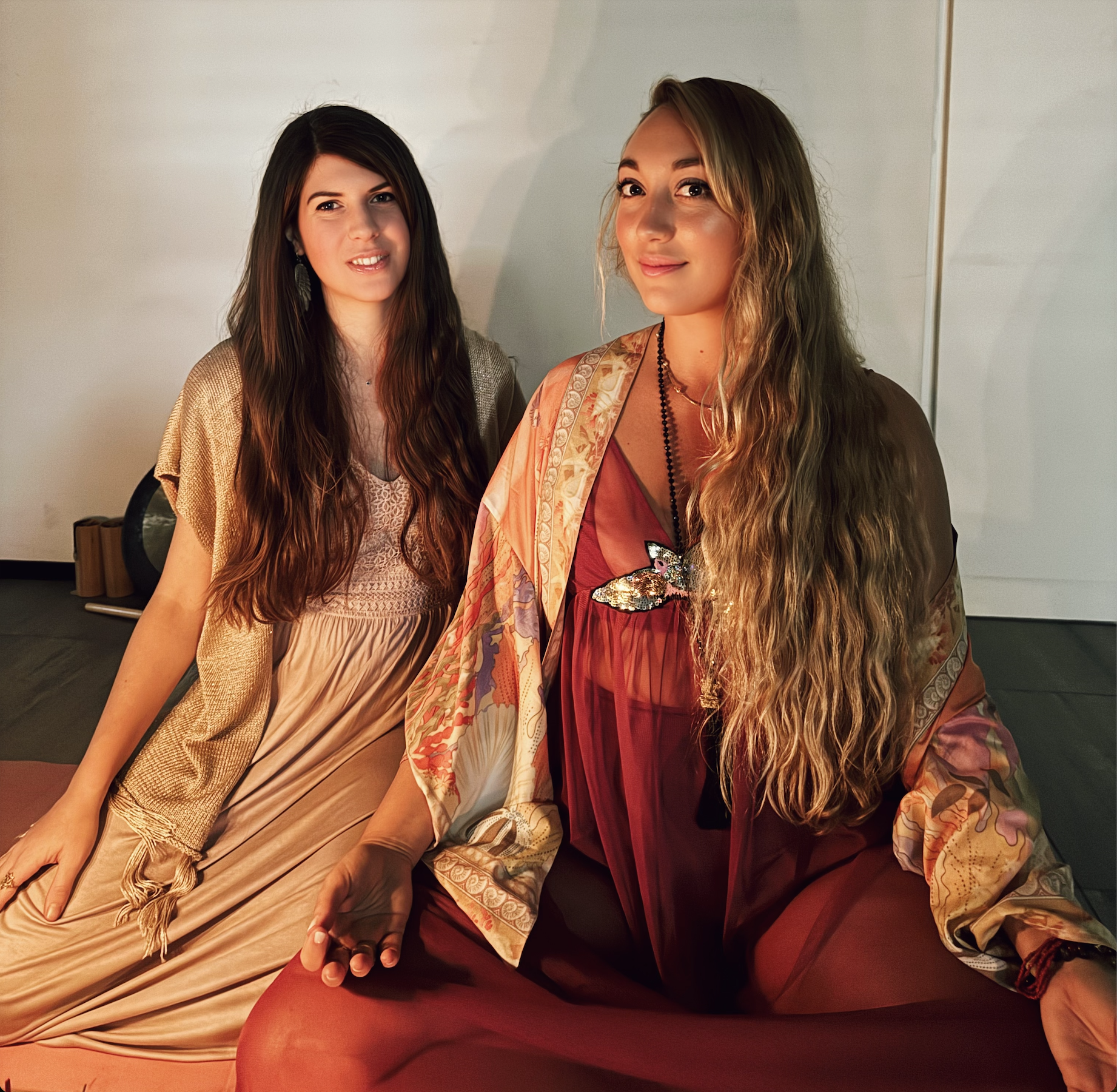 Two women sitting on the floor in a room, one with long dark hair wearing a beige dress, and the other with long curly hair wearing a red dress and layered kimono, both smiling at the camera.
