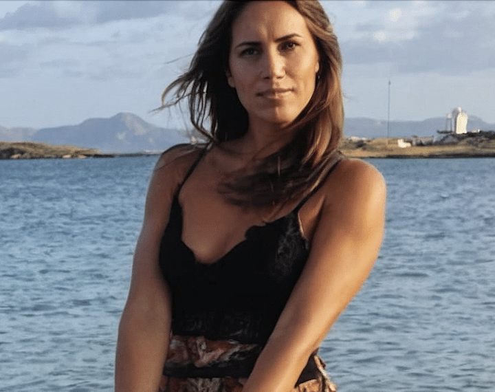 Woman with brown hair in a black sleeveless top standing near a body of water with mountains and a lighthouse in the background.