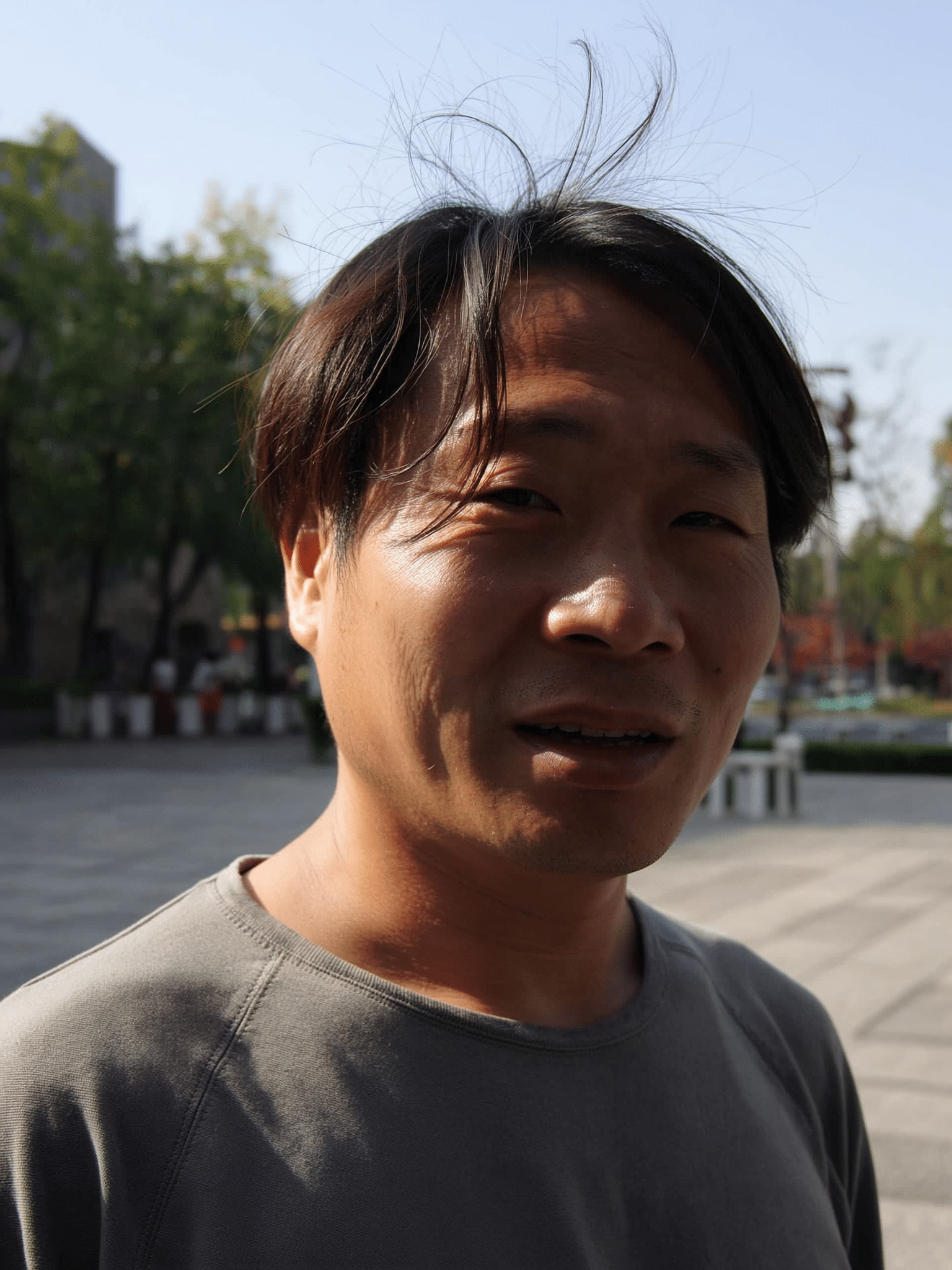 A young man with dark hair outdoors in sunlight, smiling slightly, wearing a gray shirt, with trees and a building in the background.