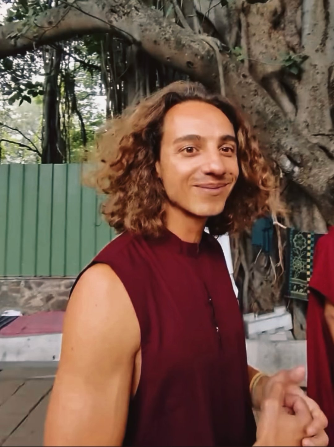 A person with curly shoulder-length hair, wearing a sleeveless maroon shirt, standing in front of a large tree with a mix of leaves and branches, and a green fence in the background.