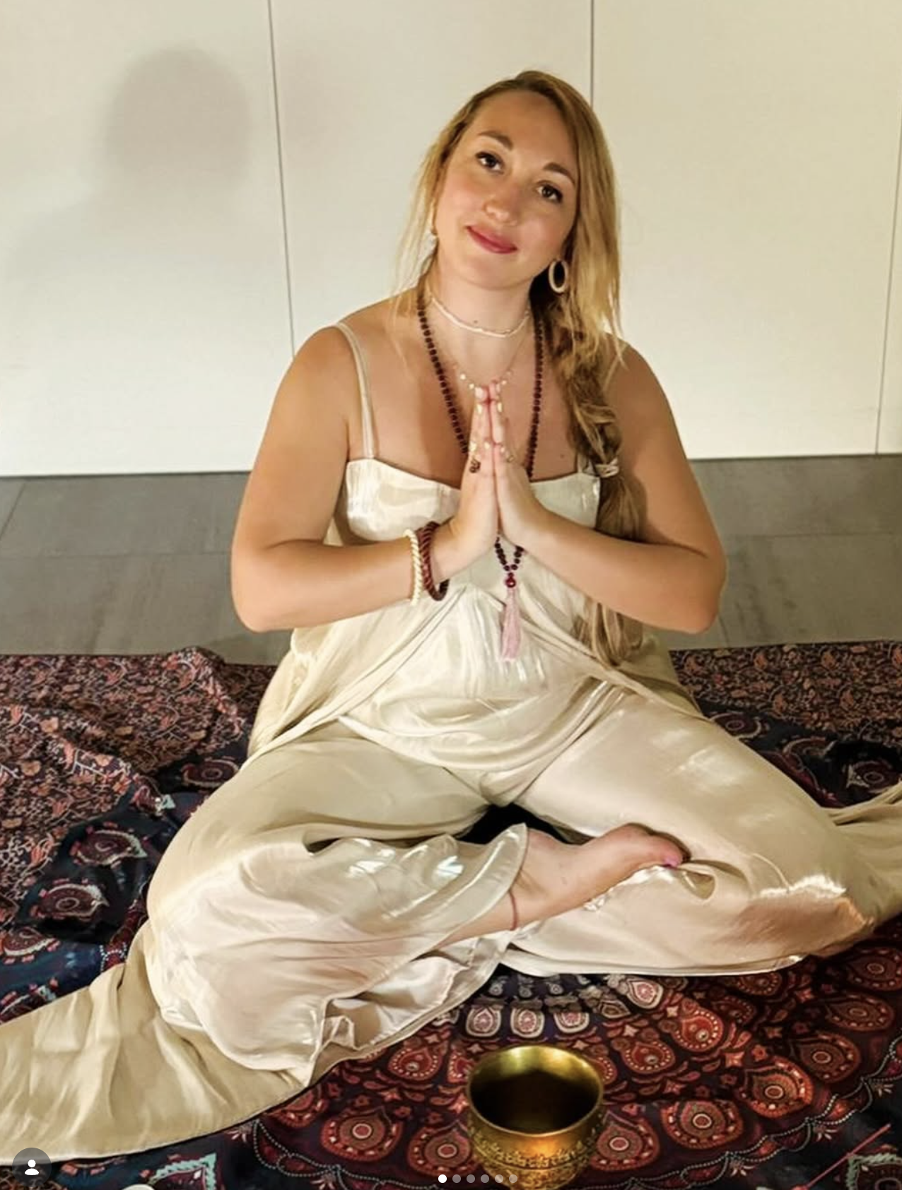A woman sitting cross-legged on a colorful patterned blanket, wearing a cream-colored satin dress, with her hands pressed together in a prayer gesture, and surrounded by spiritual jewelry including prayer beads.