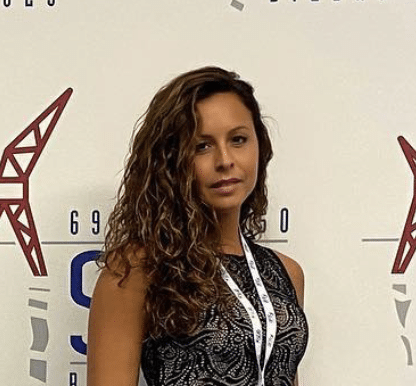 A woman with curly brown hair standing in front of a white wall with logos and text.