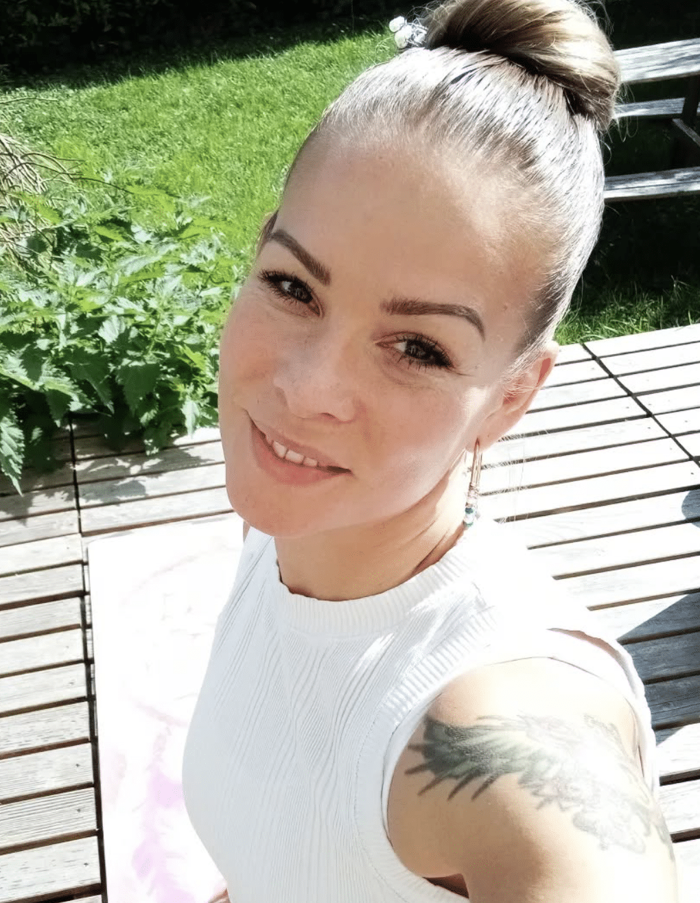 A woman taking a selfie outdoors on a sunny day, smiling, with her hair in a bun, wearing a white sleeveless top, earrings, and a tattoo on her shoulder.