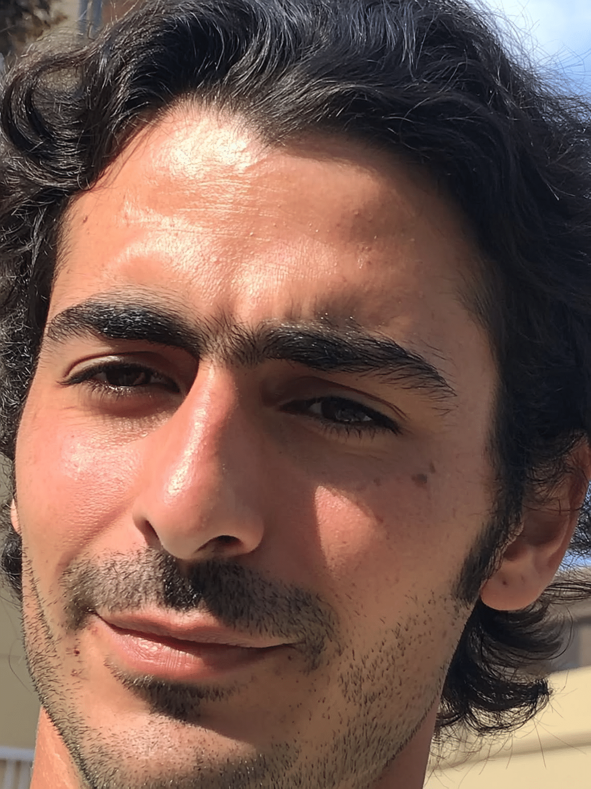 Close-up of a young man with dark, wavy hair, brown eyes, and slight facial hair, outdoors in sunlight.