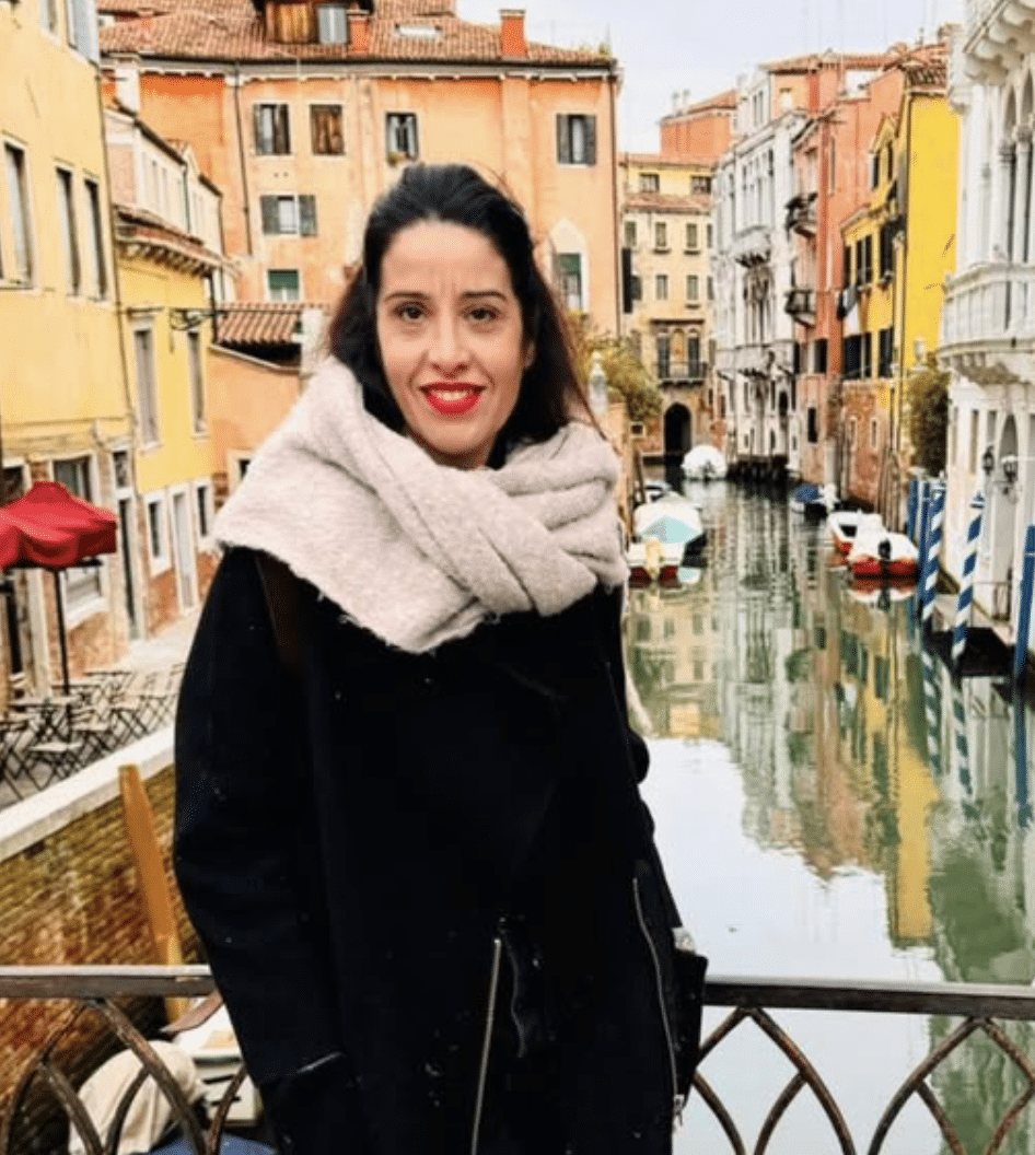 A woman standing on a bridge over a canal in Venice, Italy, with colorful buildings and boats in the background.
