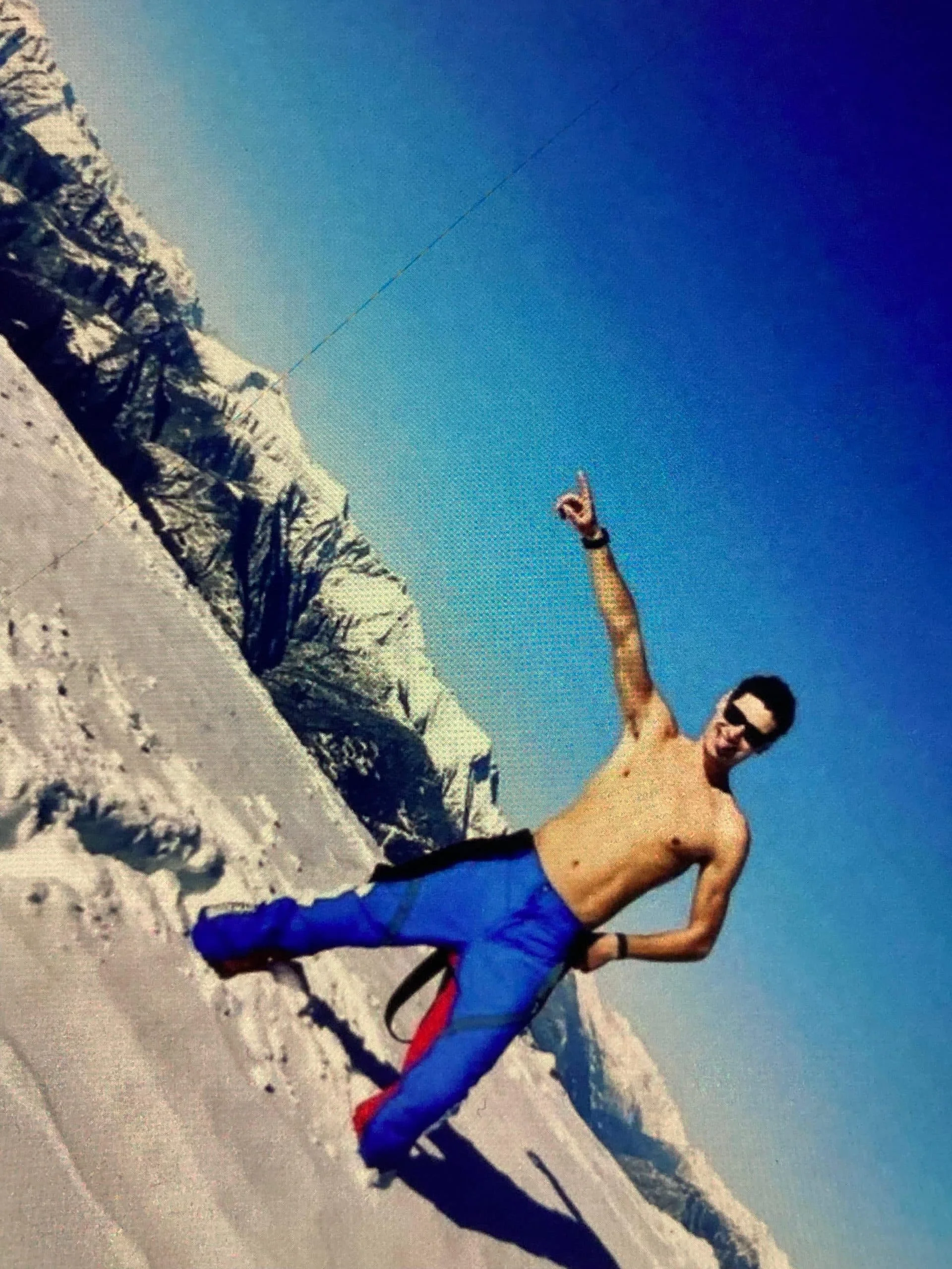 A man is shirtless, wearing blue and red snow pants, and sunglasses, standing on a snowy mountain in front of snow-covered trees and mountains, pointing upwards with his right index finger during daytime.