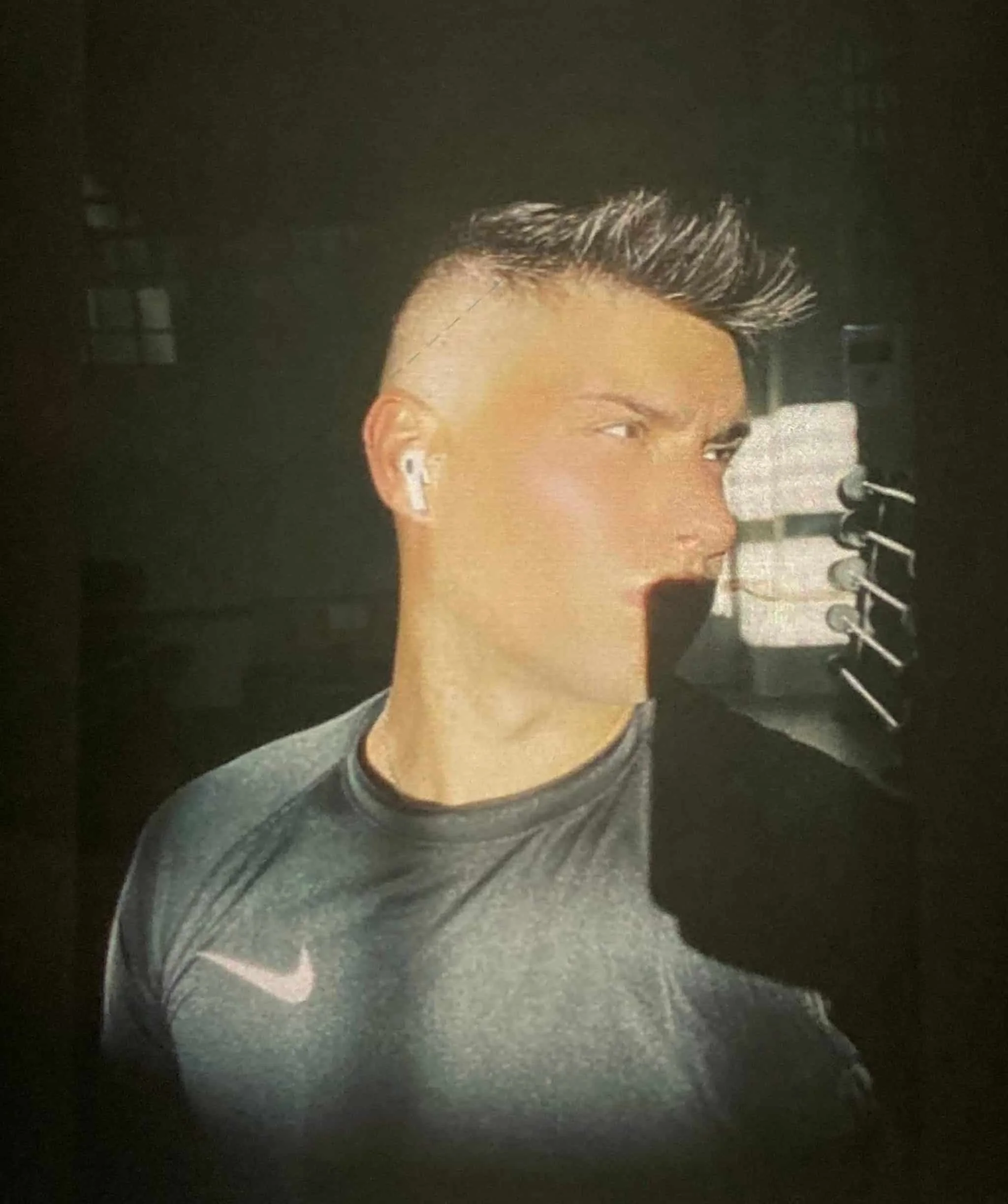 A person taking a mirror selfie in a gym, wearing a dark athletic shirt and wireless earbuds, with short hairstyle and a serious expression.