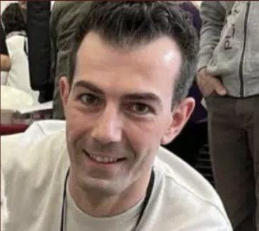 Smiling man with short, dark curly hair in a crowd, wearing a light-colored shirt.
