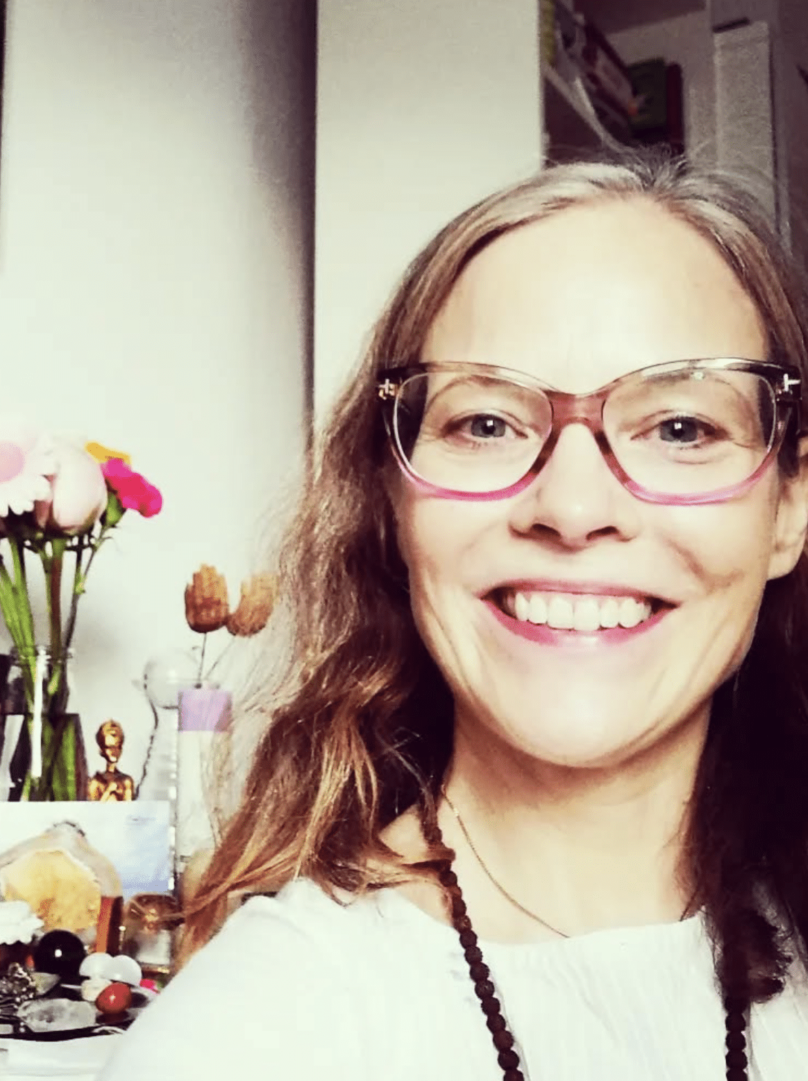 Smiling woman with long brown hair wearing glasses and a white top, standing in front of a table with various objects and a vase with pink and white flowers.