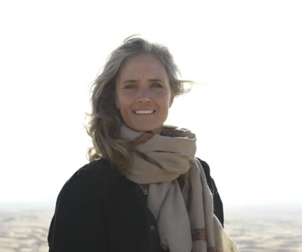 A smiling woman with gray hair wearing a black jacket and a beige scarf, outdoors on a bright day with a blurred landscape in the background.
