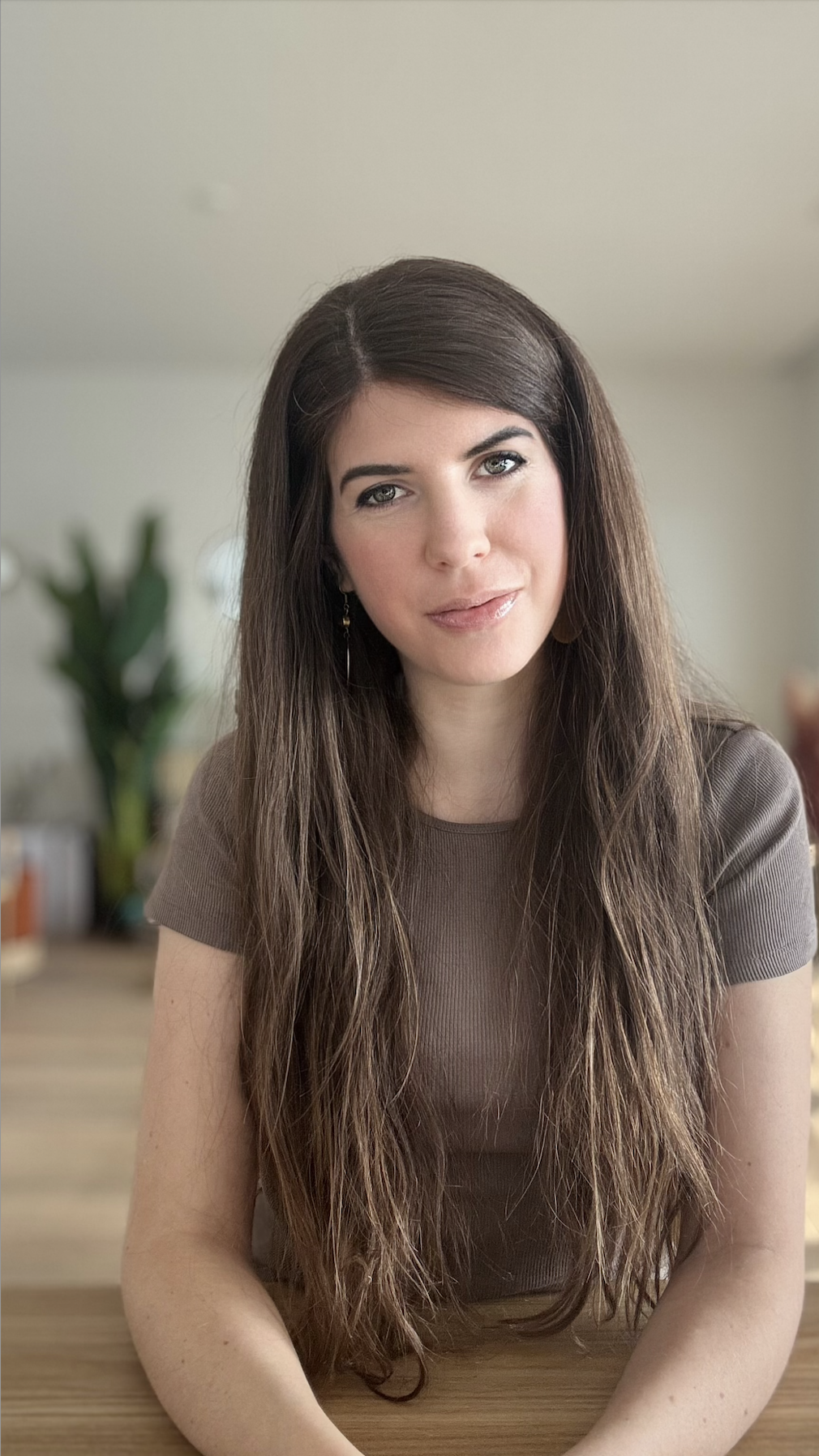 A woman with long brown hair and fair skin, wearing a brown top, sitting at a wooden table in a well-lit room with a blurred background and a large plant.