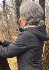 A woman with gray hair wearing a black jacket outdoors next to a tree.