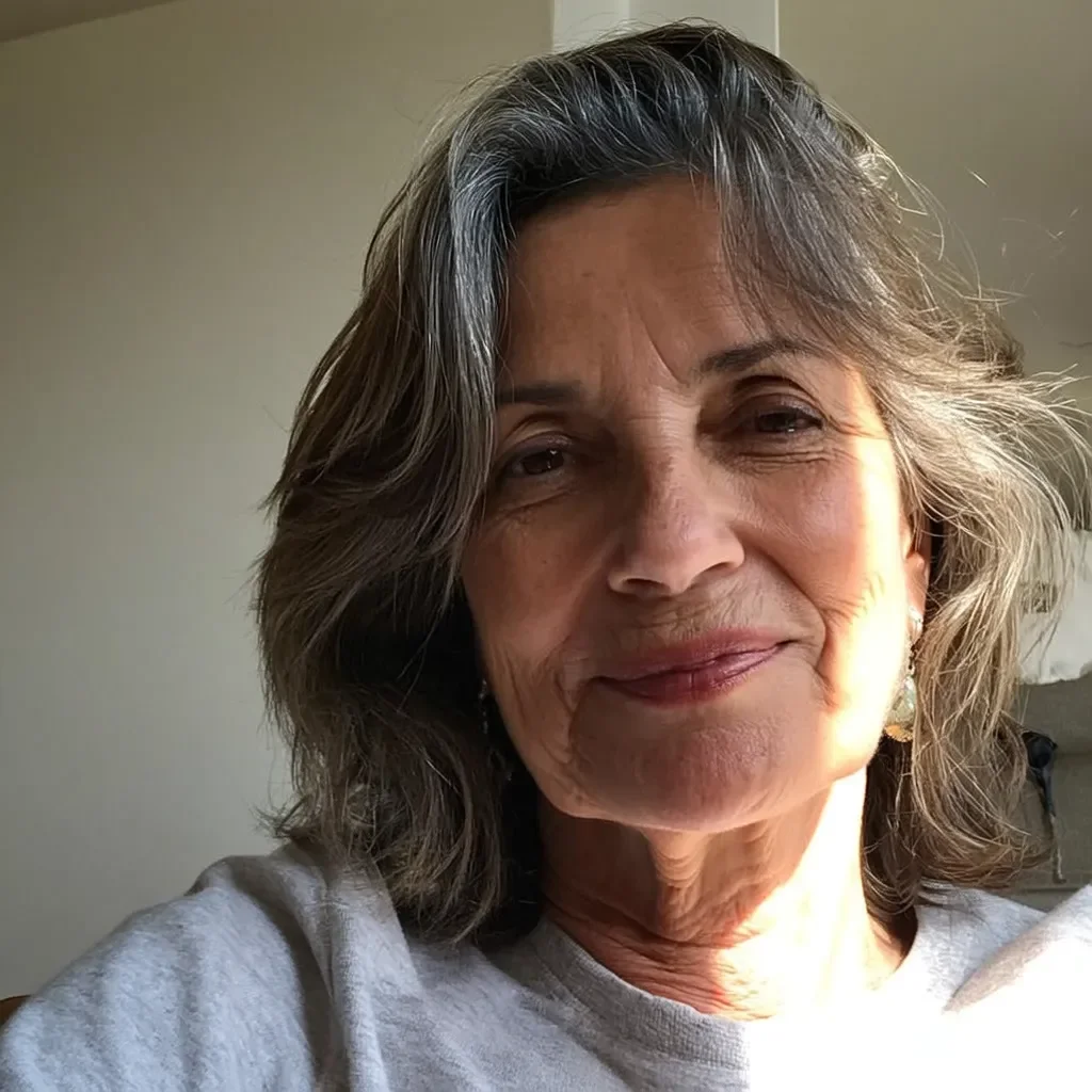 Close-up selfie of an elderly woman with gray hair and a gentle smile, wearing a light-colored top in a well-lit room.