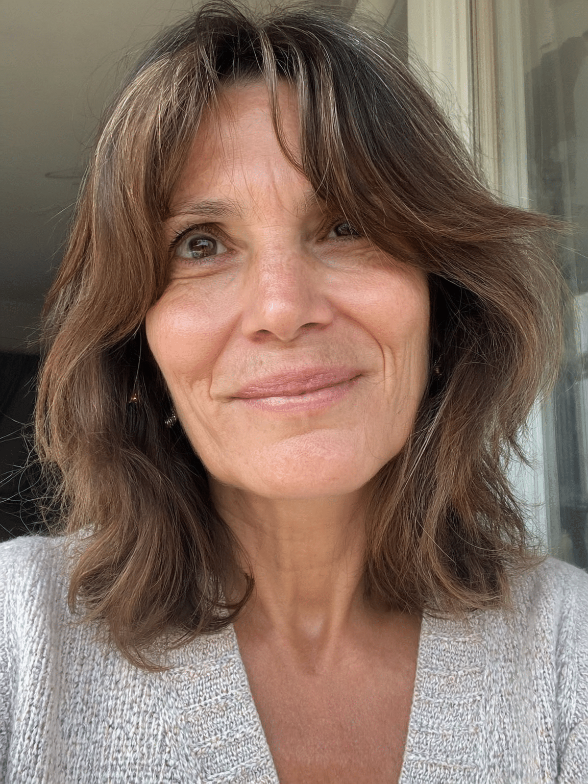A middle-aged woman with wavy brown hair and light makeup smiling at the camera indoors near a window.