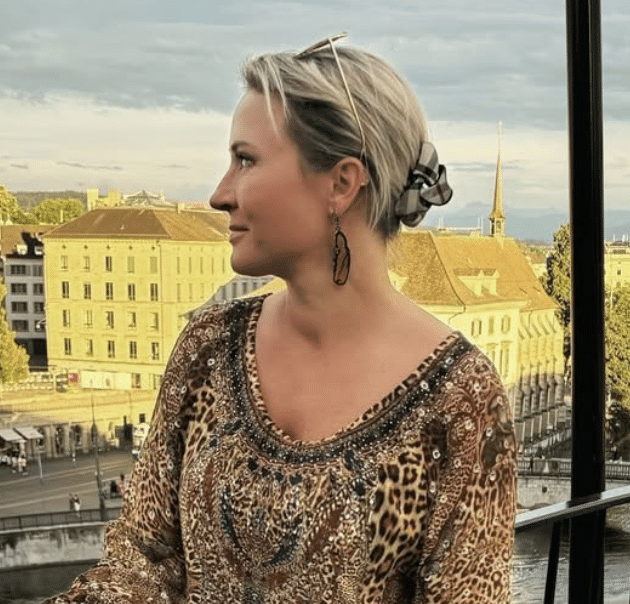 A woman with short blonde hair, wearing earrings, glasses on her head, and a multicolored animal print top, sitting on a balcony overlooking a cityscape with buildings and a church steeple.